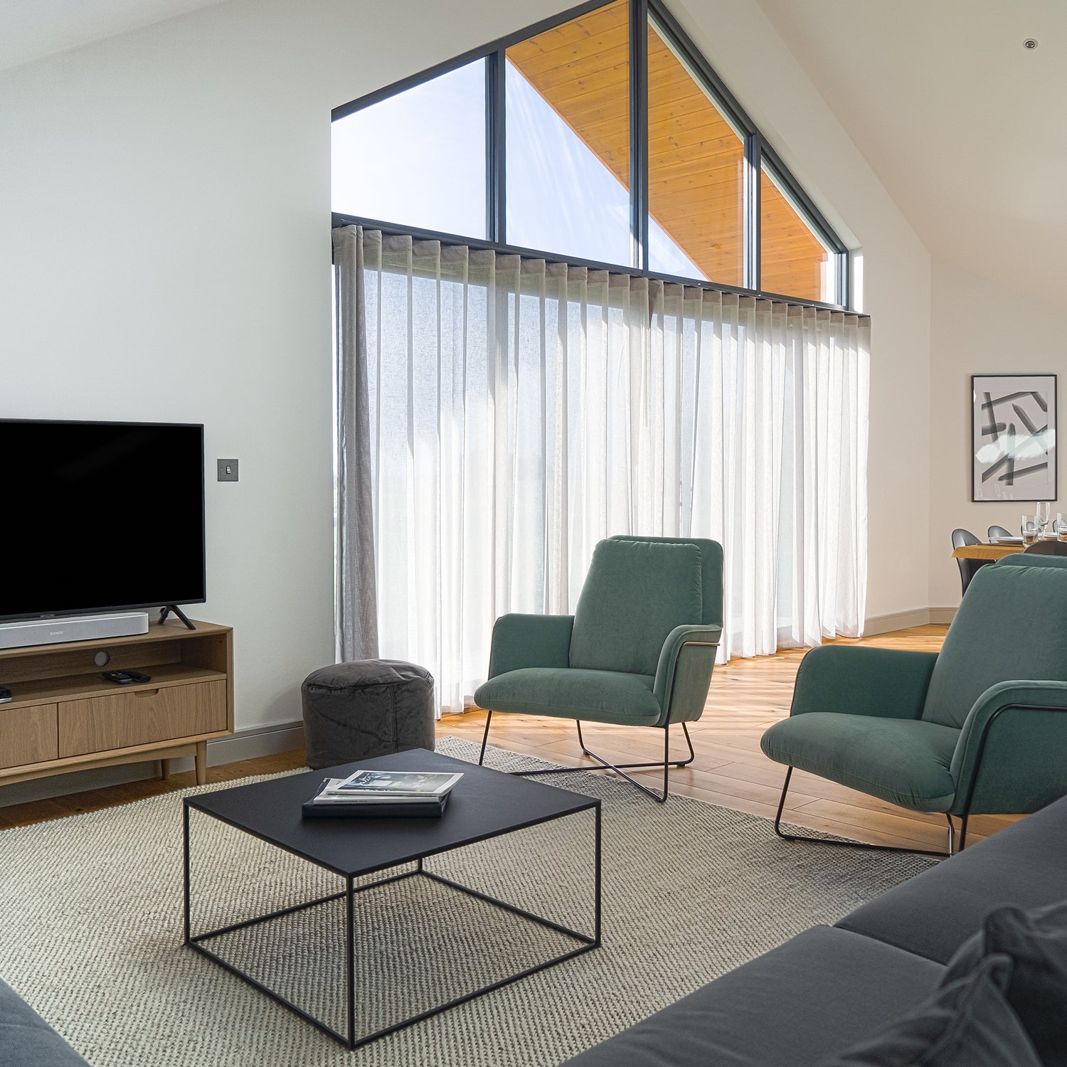 Contemporary Cornwall living room with vaulted ceiling, teal accent chairs, modern TV unit, and full-height windows with sheer curtains – bespoke interior design and stylish furniture by Iroka Interiors.