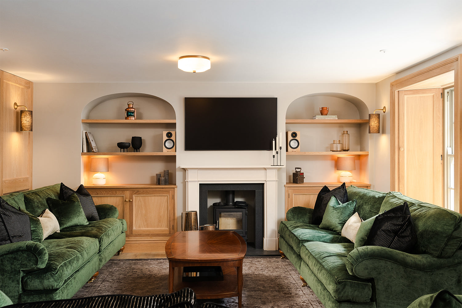 Elegant living room at Mariners, St Ives featuring deep green velvet sofas, oak built-in shelving with ambient lighting, and a central fireplace with a black wood-burning stove beneath a wall-mounted TV—designed for comfort and refined coastal charm.