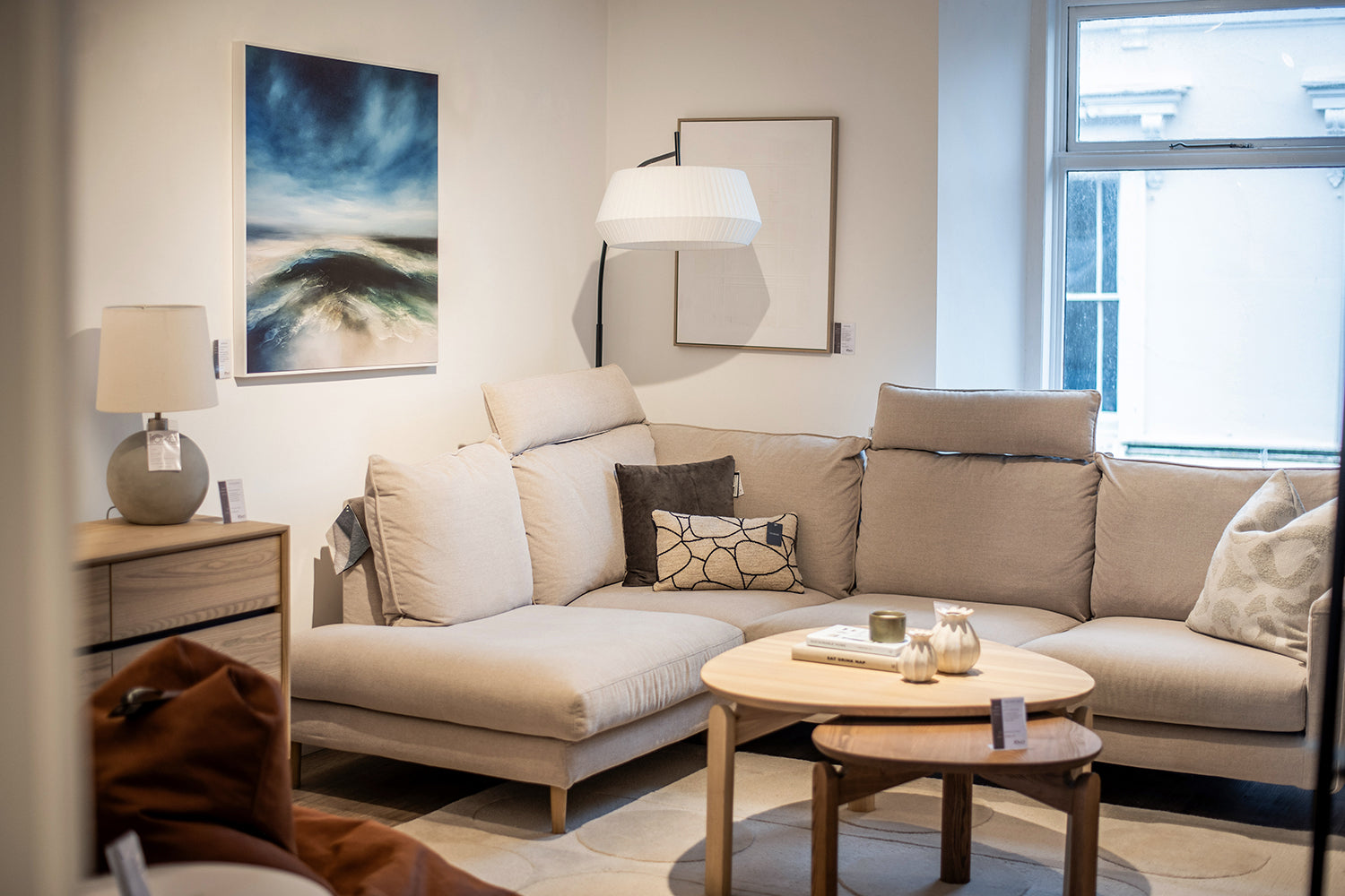 Living room set with beige corner sofa and wooden natural accents. Blue seascape canvas on the wall and neutral coloured soft furnishings.