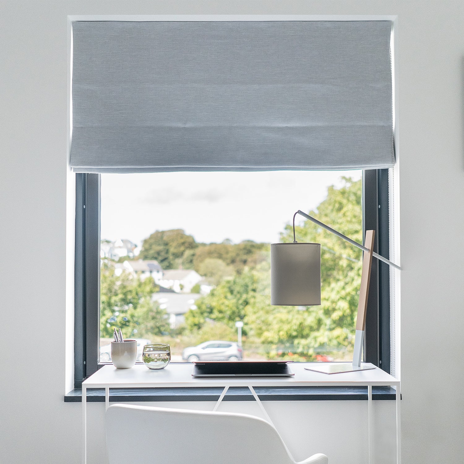Tailored Roman blind in soft grey fabric, fitted in a modern home office with garden views — bespoke blinds by luxury interior design and window dressing specialist.