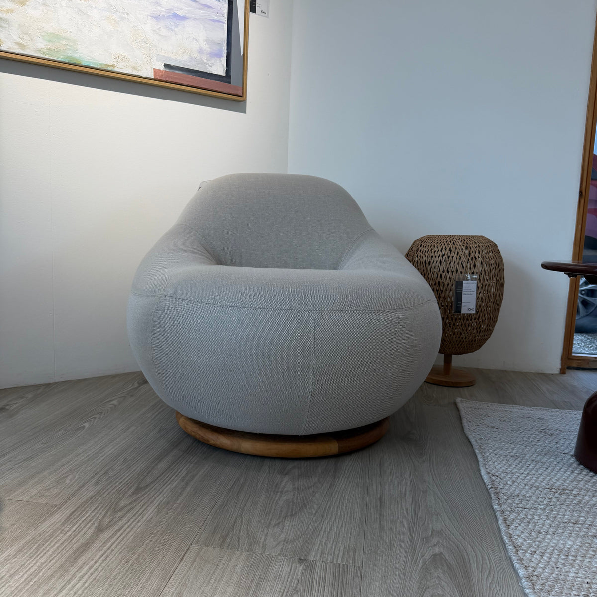 A modern round swivel armchair upholstered in light gray textured fabric with a natural wood base, shown in a minimalist room setting with gray wood-look flooring and a framed painting on the wall.
