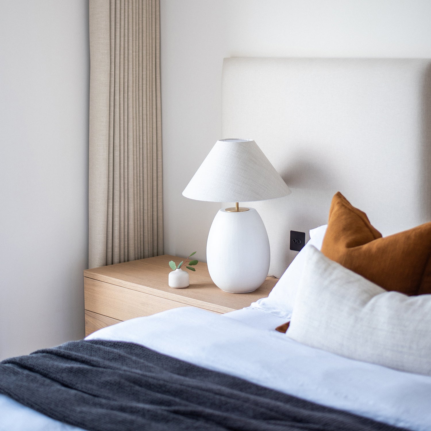 Close-up of modern bedroom with neutral upholstered headboard, white ceramic table lamp, oak bedside table, layered linen bedding, and rust accent cushions – minimalist and cozy interior styling