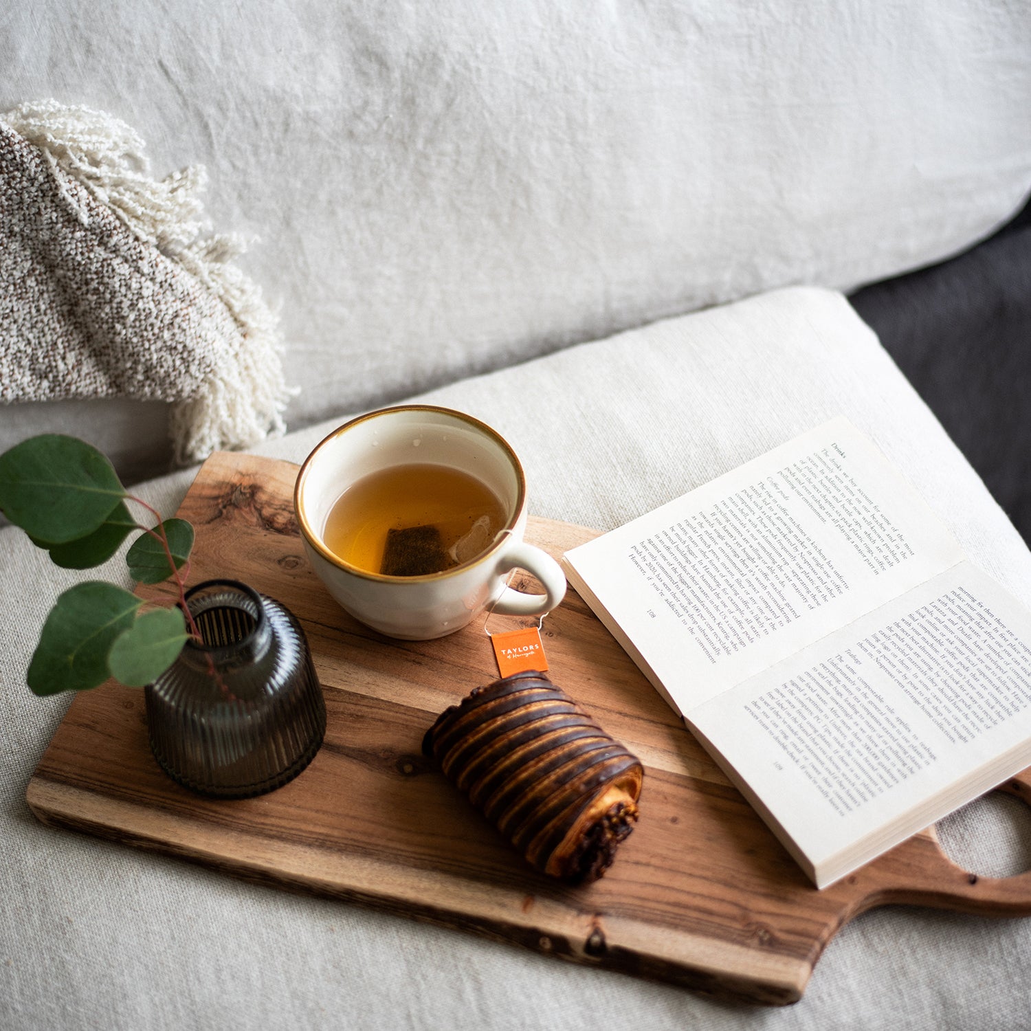 Relaxing setup with a cup of tea, a pain au chocolat, an open book, and a small vase with greenery on a wooden serving board, resting on a soft, neutral-toned sofa.”