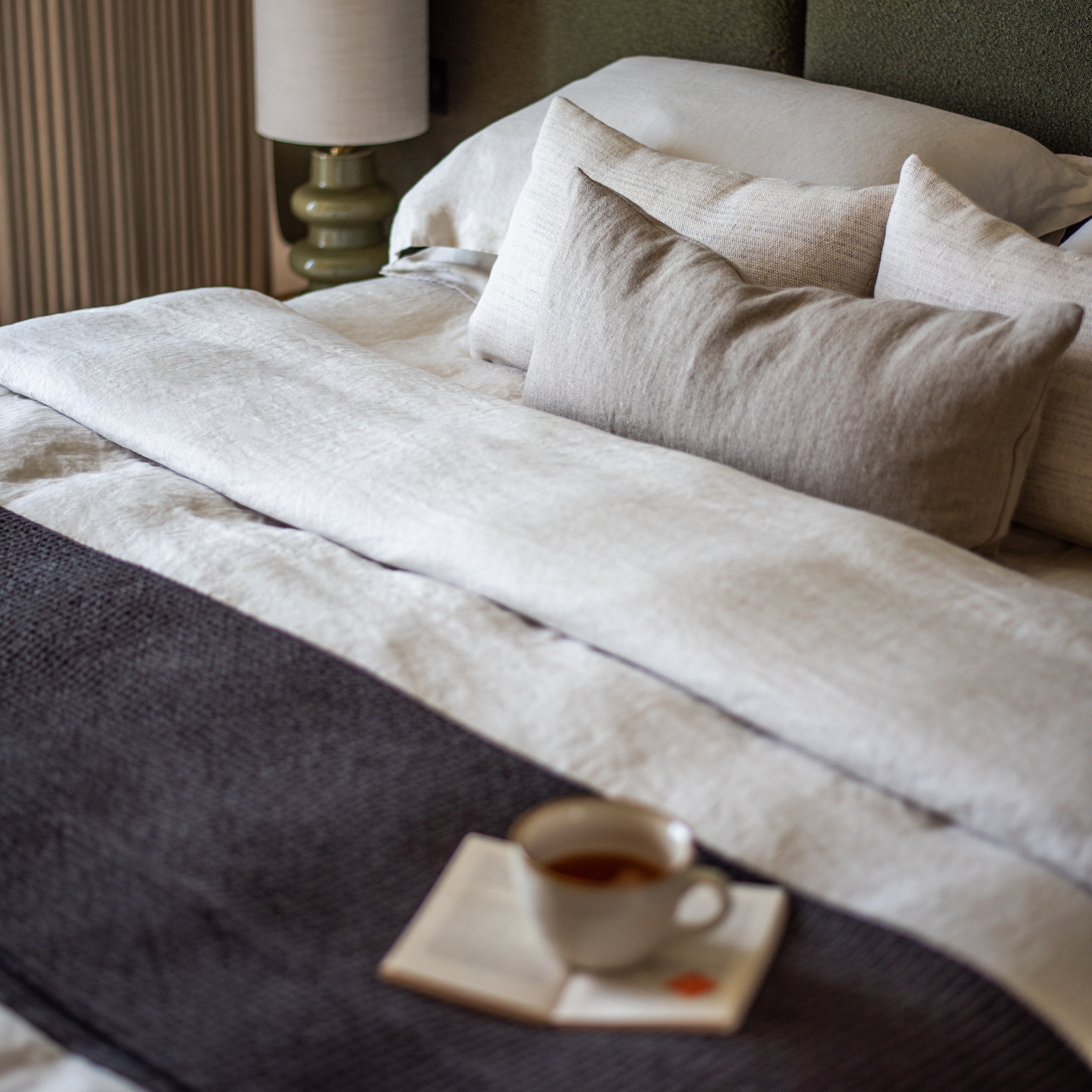 Luxurious bed with layered natural linen bedding, soft neutral cushions and textured throw, featuring a morning cup of tea and open book – serene bedroom styling at The Cliffs, Carbis Bay.