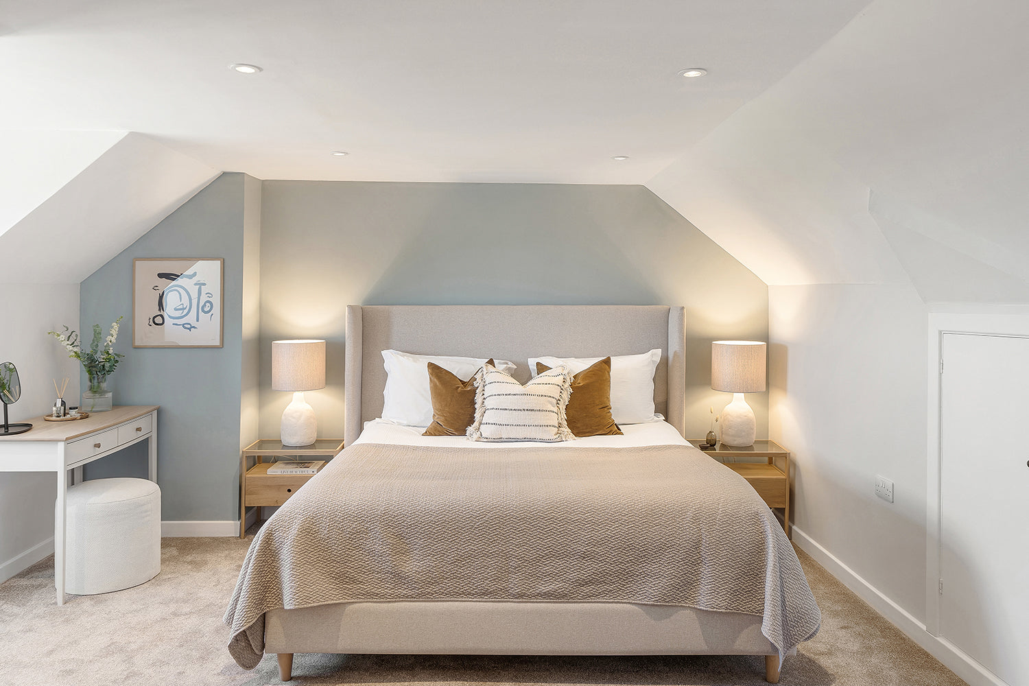 Light and airy attic bedroom at Fairskies, Merryn featuring a soft grey upholstered bed, neutral layered bedding, warm ochre cushions, ceramic bedside lamps, and a minimalist vanity with abstract artwork – styled by Iroka Interiors.