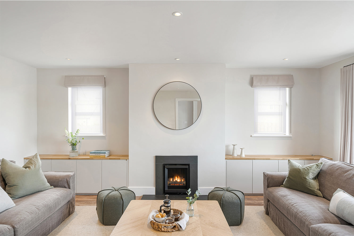 Scandinavian-style living room at Fairskies, Merryn featuring symmetrical sofas, a modern wood-burning stove, round wall mirror, neutral palette with sage green accents, and minimalist built-in cabinetry – interior design by Iroka.