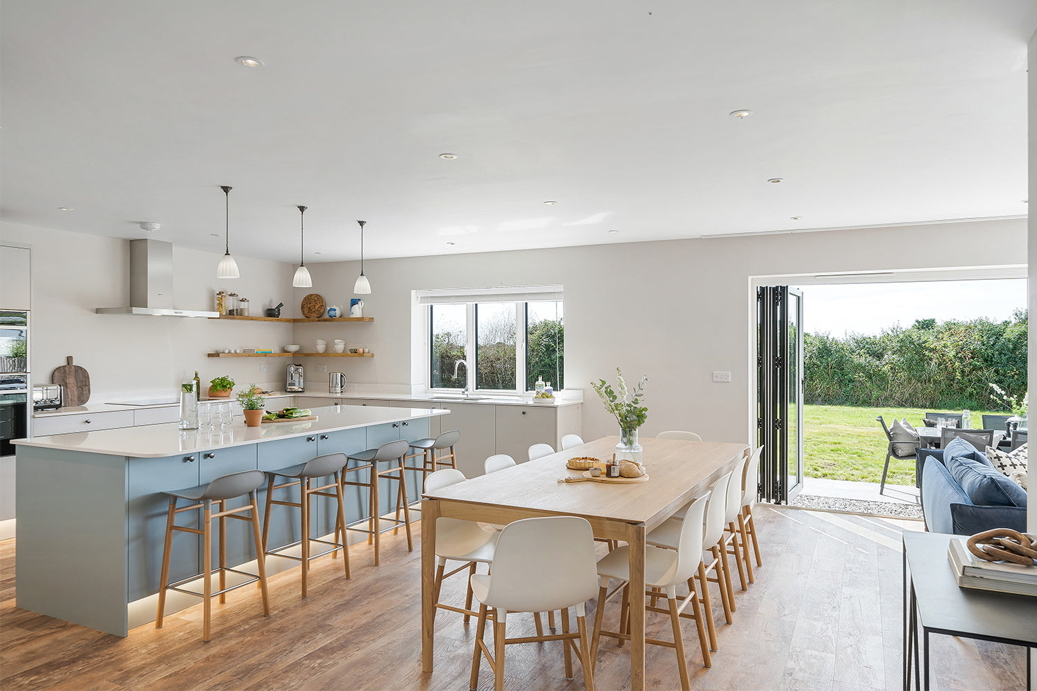 Open-plan modern farmhouse kitchen and dining area in Fairskies, Merryn – features light wood flooring, blue bar stools, quartz waterfall island, open oak shelving, and panoramic sliding doors with countryside views – styled by Iroka Interiors.