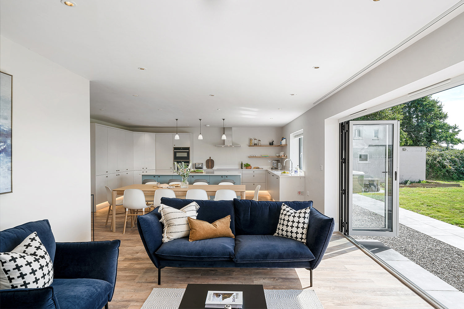 Contemporary open-plan kitchen, dining, and living space in Fairskies, Merryn – navy velvet sofas, minimalist white cabinetry, light wood flooring, and bi-fold doors opening to a landscaped garden – designed by Iroka Interiors.