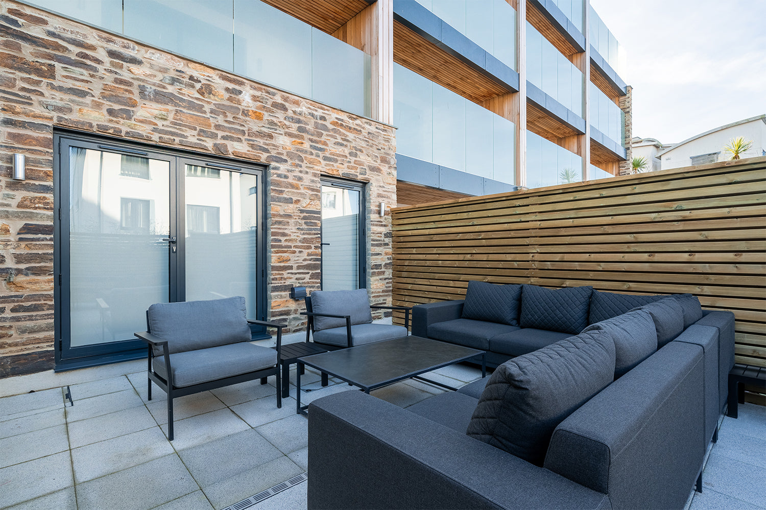 Modern outdoor patio area featuring dark grey upholstered lounge furniture, black metal frame coffee table, and contemporary stone and timber exterior walls at a stylish apartment complex.