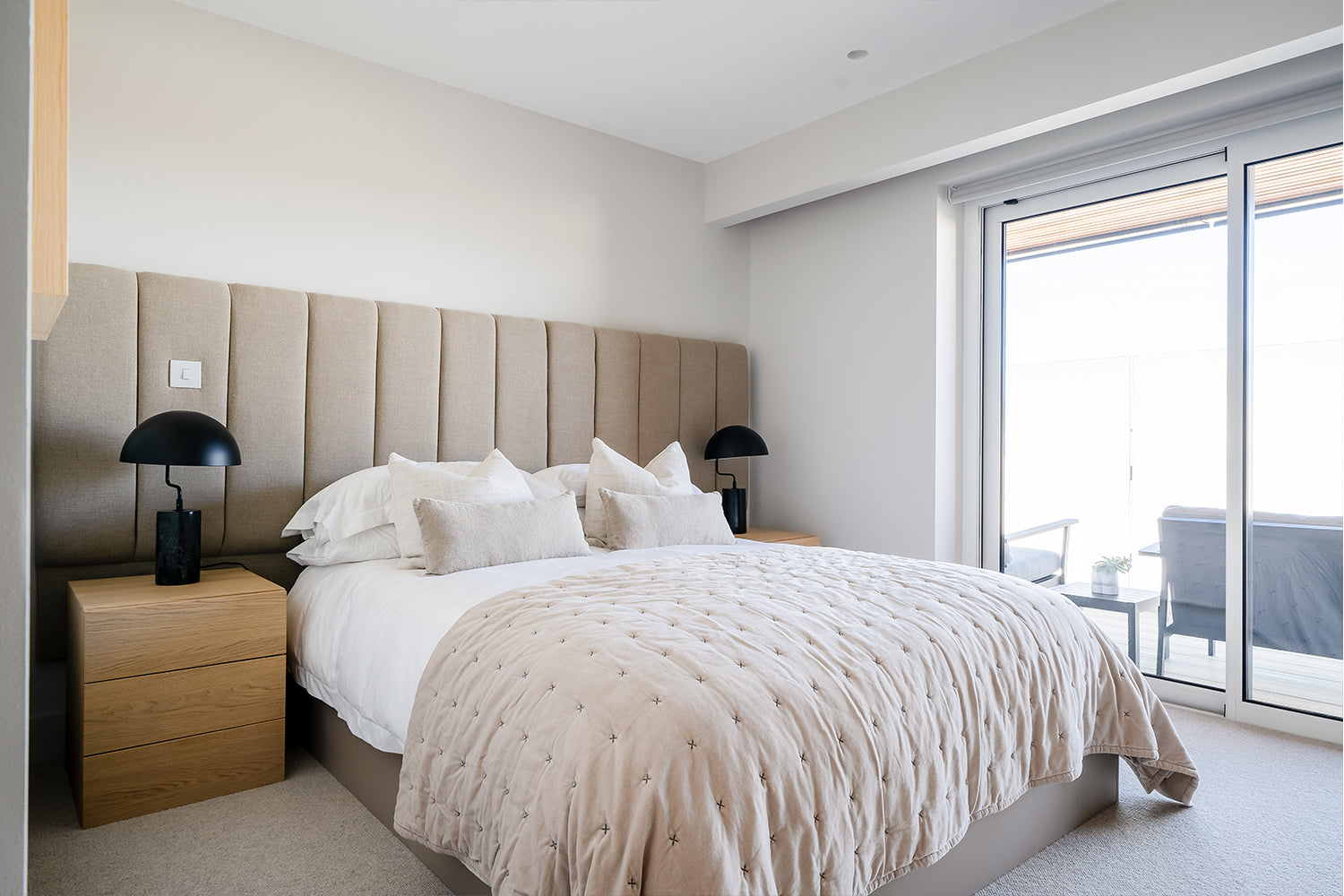Contemporary coastal bedroom with a beige upholstered headboard, crisp white bedding, and neutral cushions. Natural wood bedside tables and black modern lamps enhance the minimalist decor, with sliding glass doors opening to a private balcony.