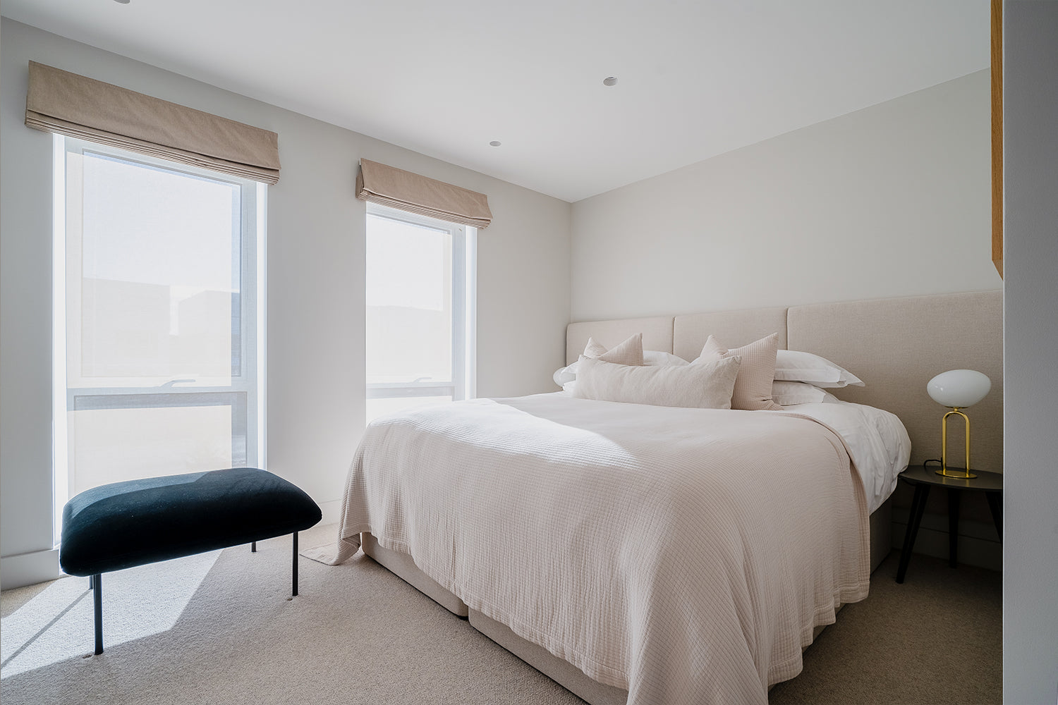 Bright minimalist bedroom with soft neutral tones, featuring a king-sized bed with beige headboard and layered cushions, two large windows with taupe Roman blinds, a black bench, and a modern white and gold bedside lamp.