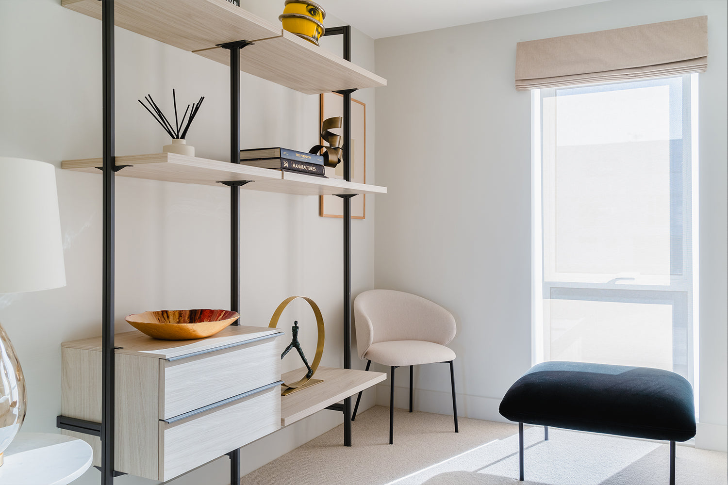 Modern home office corner with open wooden shelving, decorative accessories, sculptural ornaments, and neutral furnishings including a cream accent chair and black upholstered bench by a large window with Roman blinds.
