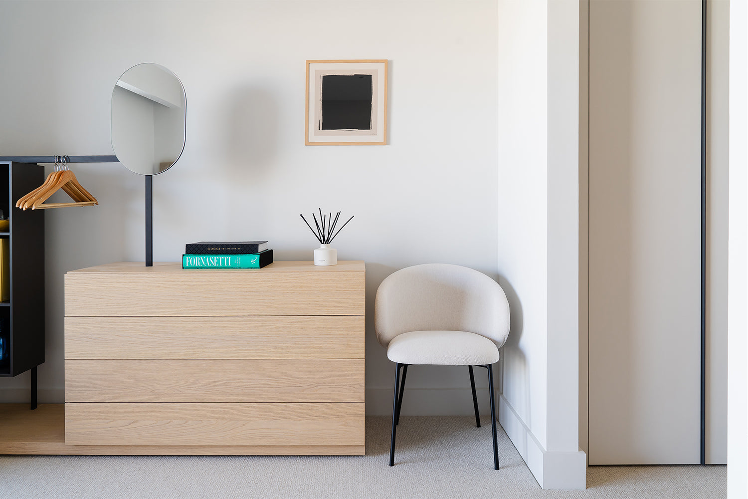 Minimalist bedroom corner featuring a light oak chest of drawers with books and a diffuser, a modern vanity mirror, a rounded cream chair with black legs, and a small wall-mounted artwork.
