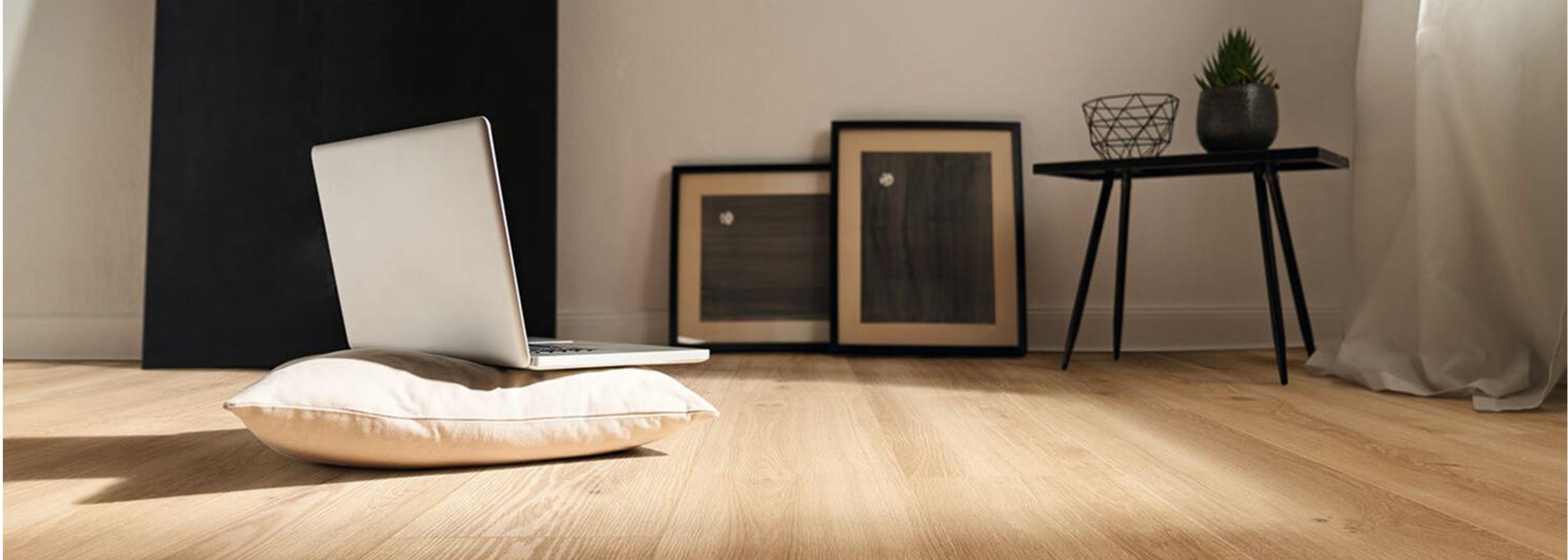 Modern minimalist room featuring natural oak-look luxury vinyl tile flooring, with a laptop on a cushion and framed artwork leaning against a wall, conveying a warm and contemporary home office ambiance.