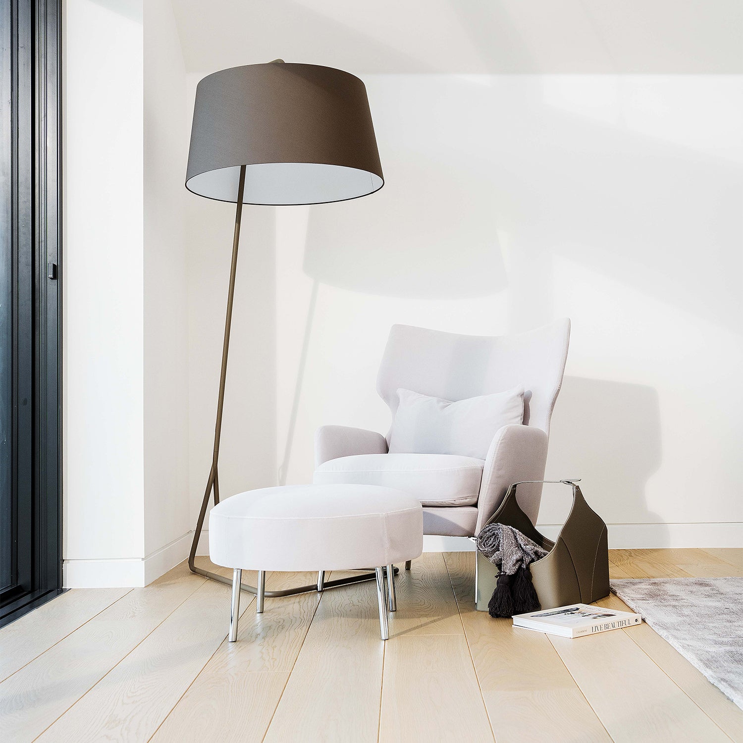 Light-toned engineered wood flooring in a modern minimalist living room featuring a white armchair, ottoman, floor lamp, and soft natural lighting from sliding glass doors.