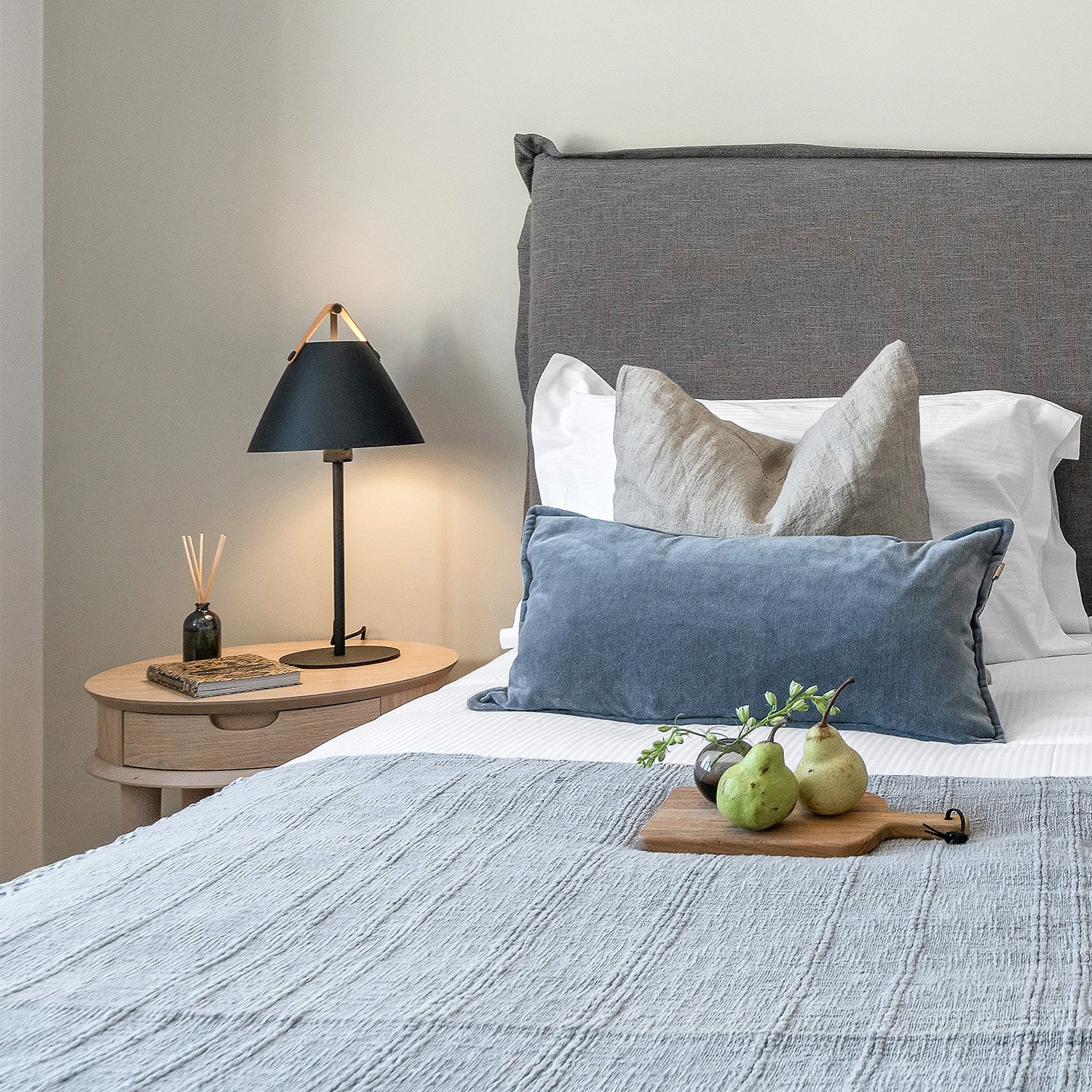 Cozy modern bedroom with charcoal upholstered headboard, layered linen and velvet cushions, wooden bedside table, black designer lamp, and styled bed with textured throw – serene interior design with soft neutral palette.