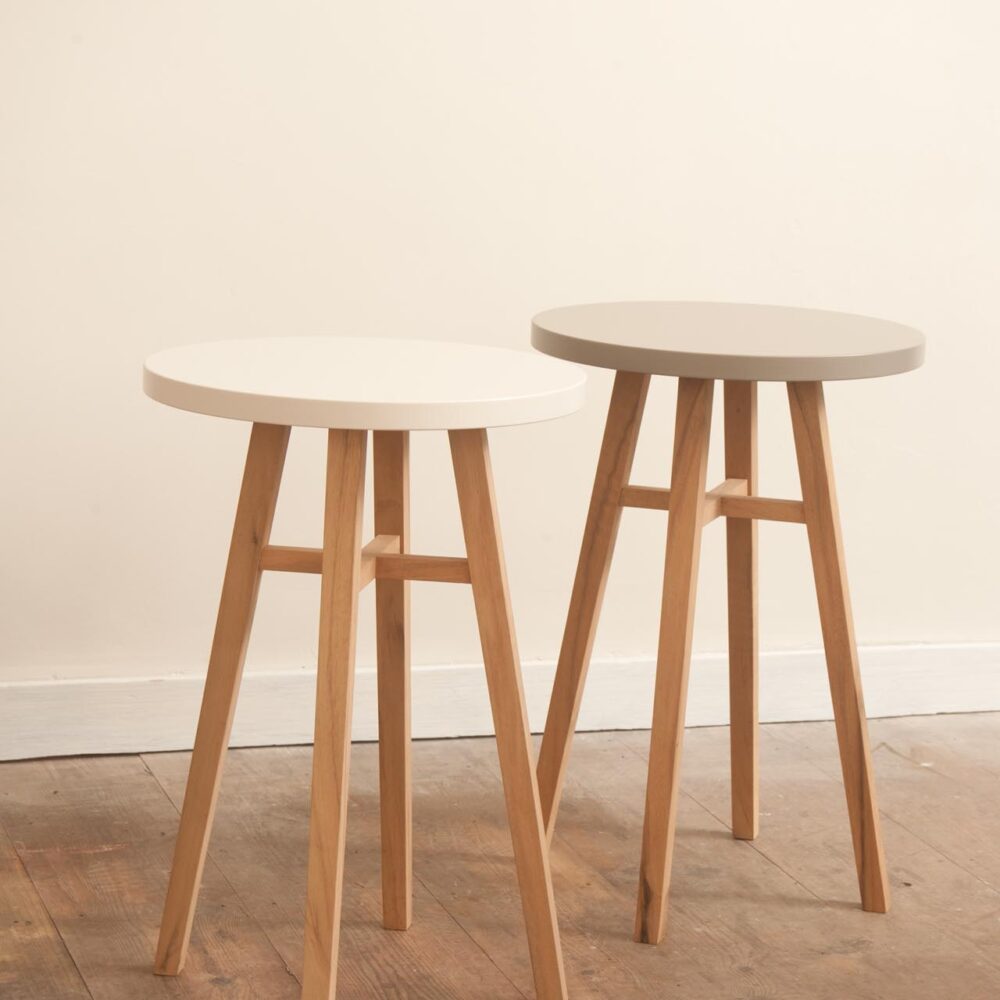 Two round nesting side stools with light natural wood legs and one stool featuring a white top and the other a light grey top, set against a plain cream wall and wooden floor.