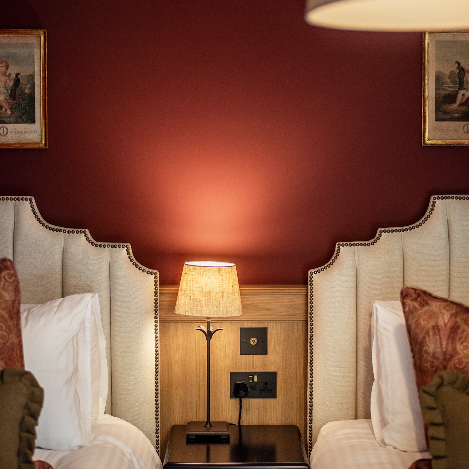 Classic twin bedroom with scalloped upholstered headboards, warm burgundy feature wall, antique-style artwork, wooden paneling, and ambient table lamp – traditional luxury interior design with rich tones.