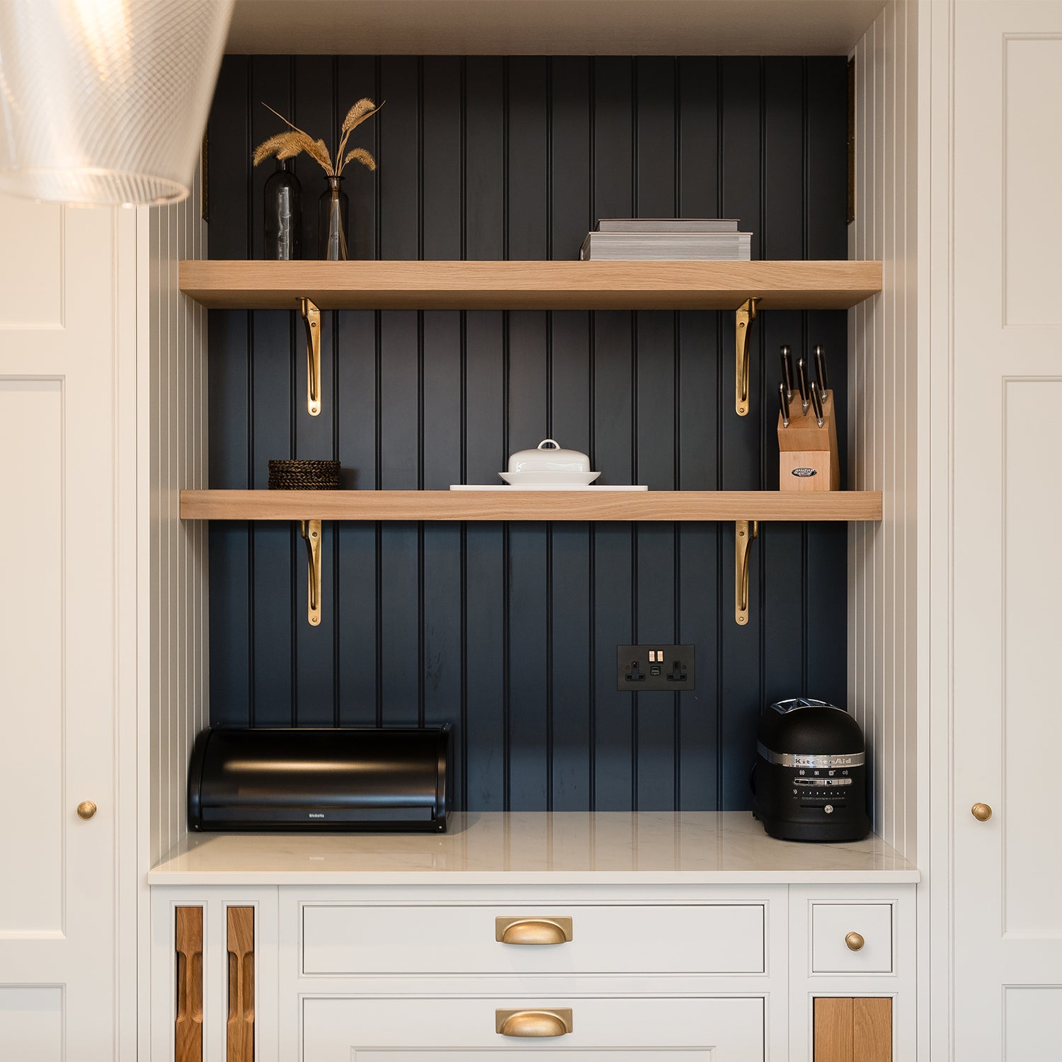 Bespoke kitchen nook at Mariners, St Ives featuring integrated white cabinetry with brass handles, deep navy tongue-and-groove panelled back, floating oak shelves, and elegant brass brackets—styled with accessories and built-in appliances.