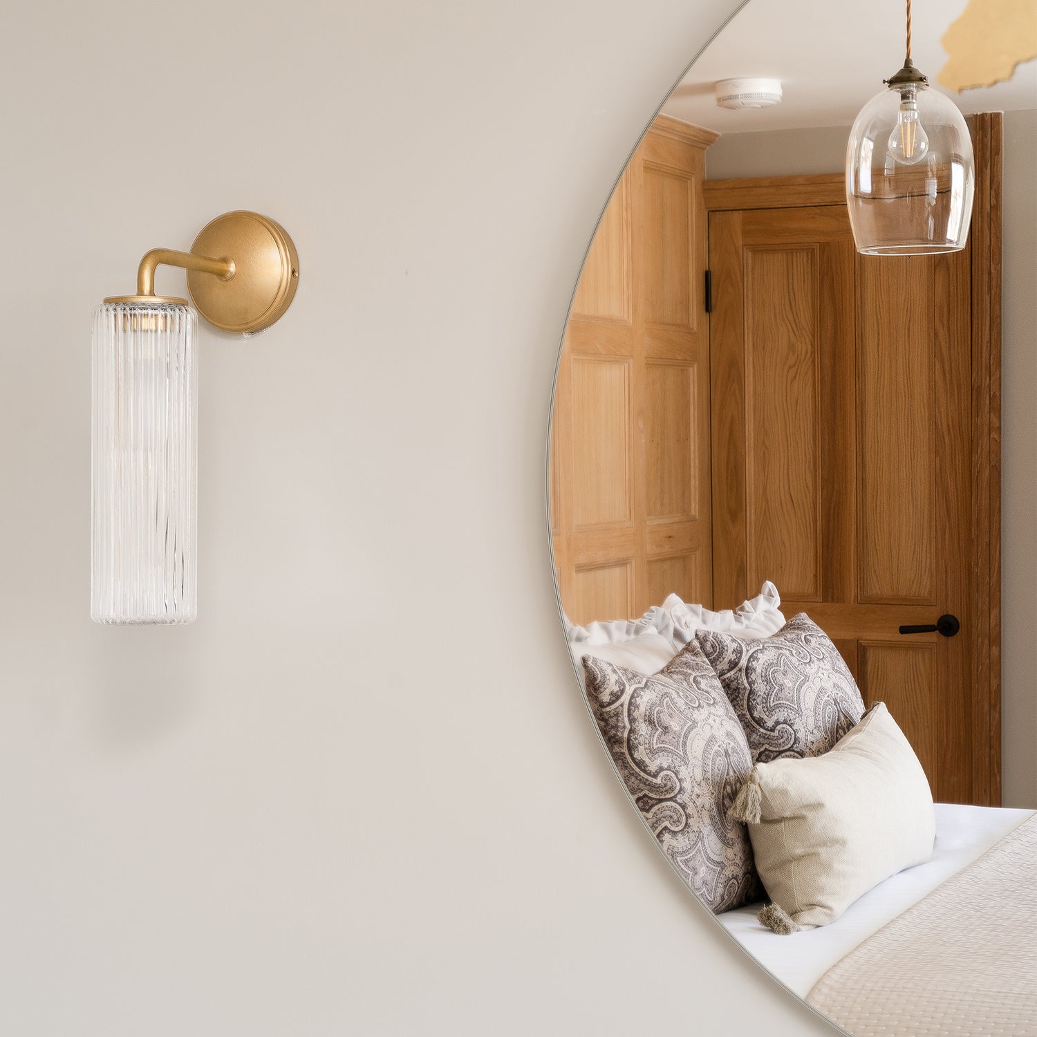 Detail of a round wall mirror reflecting a coastal bedroom at Mariners, St Ives, with oak panelled doors, ruffled paisley cushions, and a neutral palette. Brass wall light and glass pendant add a touch of elegance to the soft, contemporary interior.