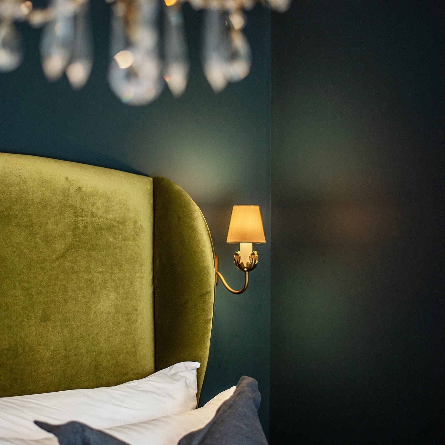 Close-up of a luxurious olive green velvet headboard set against deep teal walls in a bedroom at Mariners, St Ives. A brass wall sconce with a warm shade and crystal chandelier details create a rich, elegant ambience.