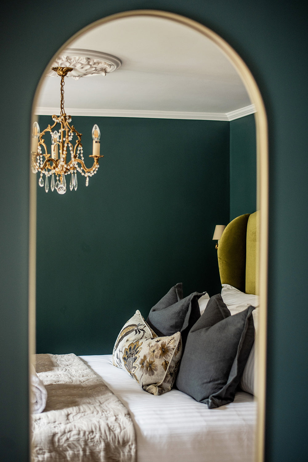Refined bedroom at Mariners, St Ives, featuring a velvet olive green headboard, dark teal walls, ornate gold chandelier, and layered cushions in rich, muted tones viewed through an arched mirror.