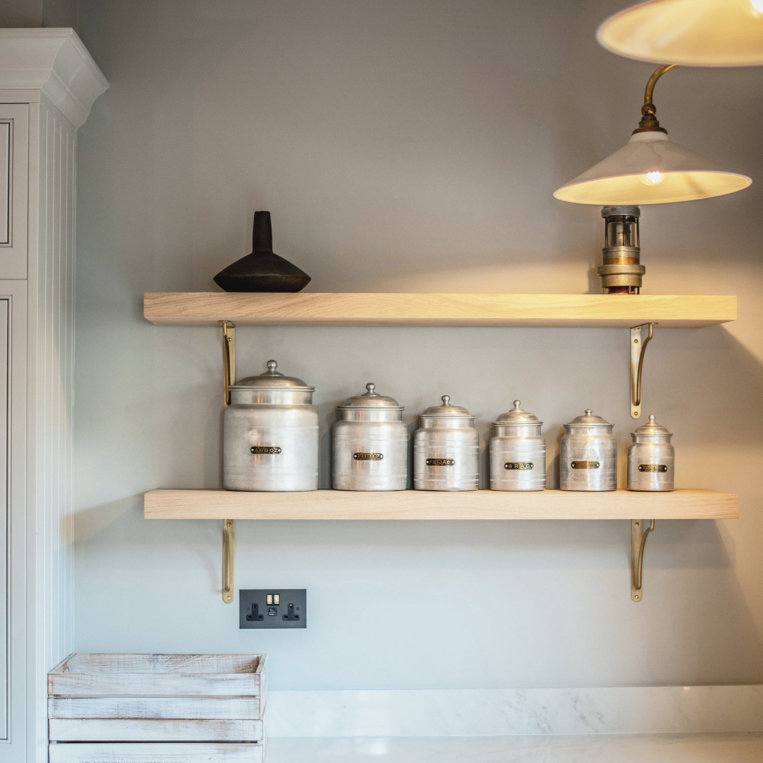 Open kitchen shelving at Mariners, St Ives featuring natural oak shelves with brass brackets, a vintage-style ceramic canister set, and pendant lighting with fluted glass shades—set against a soft grey wall for a modern coastal finish.