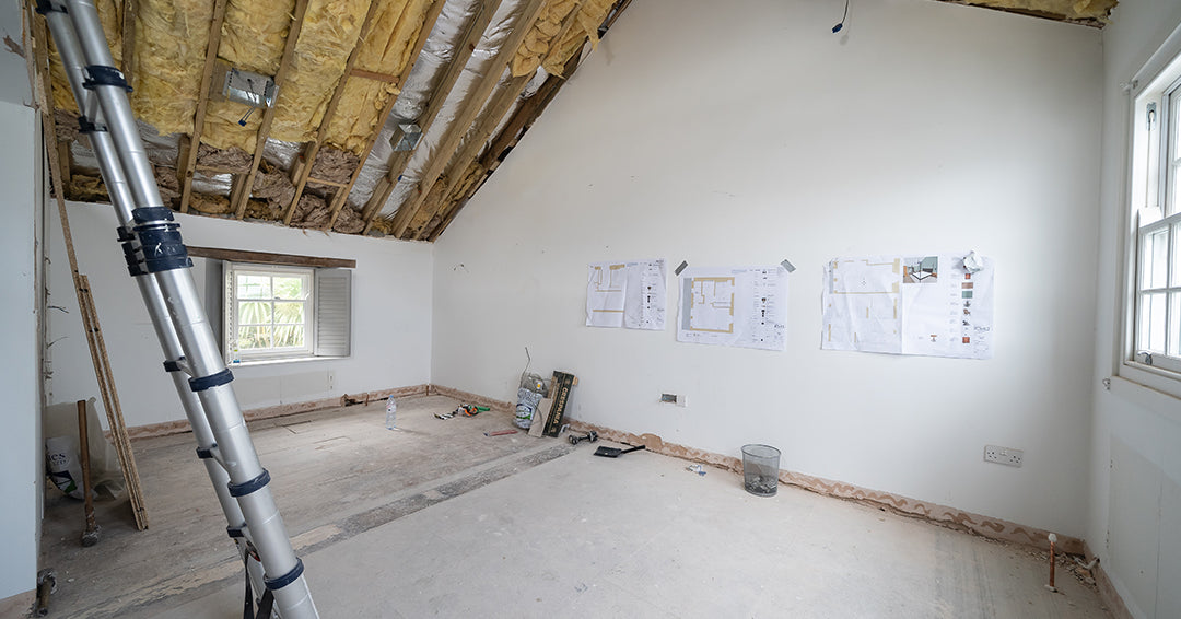 Master bedroom renovation in progress with exposed insulation, partially stripped flooring, architectural plans on wall, and natural light from double-hung windows – interior remodeling and conversion before image.
