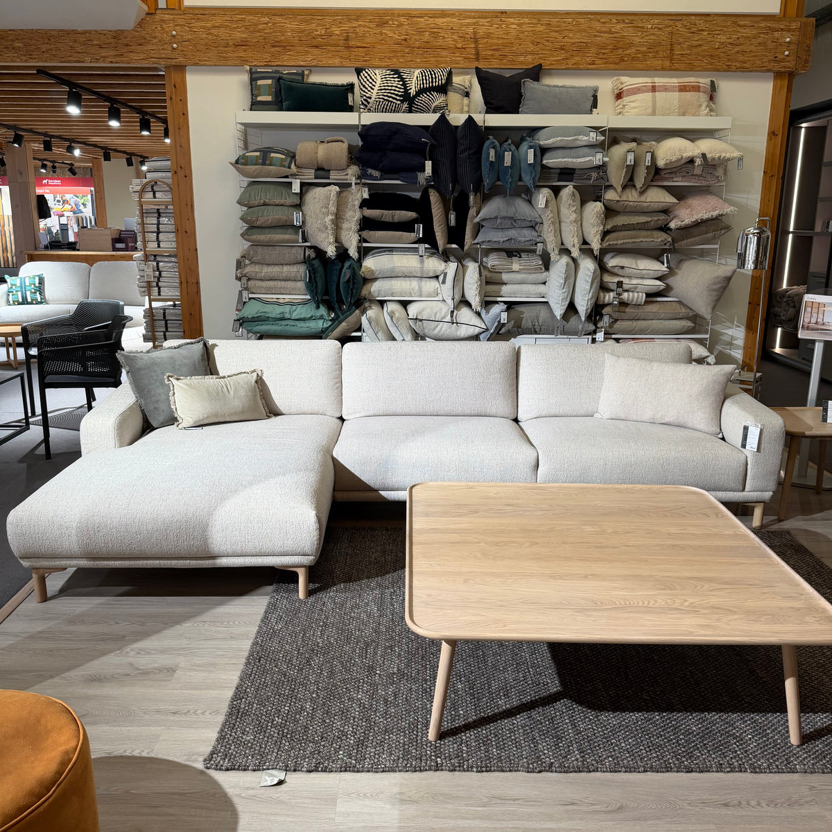 A light gray modular sectional sofa with a chaise lounge, styled in a furniture showroom with a light wood coffee table, dark gray rug, and a wall of folded textiles behind it.