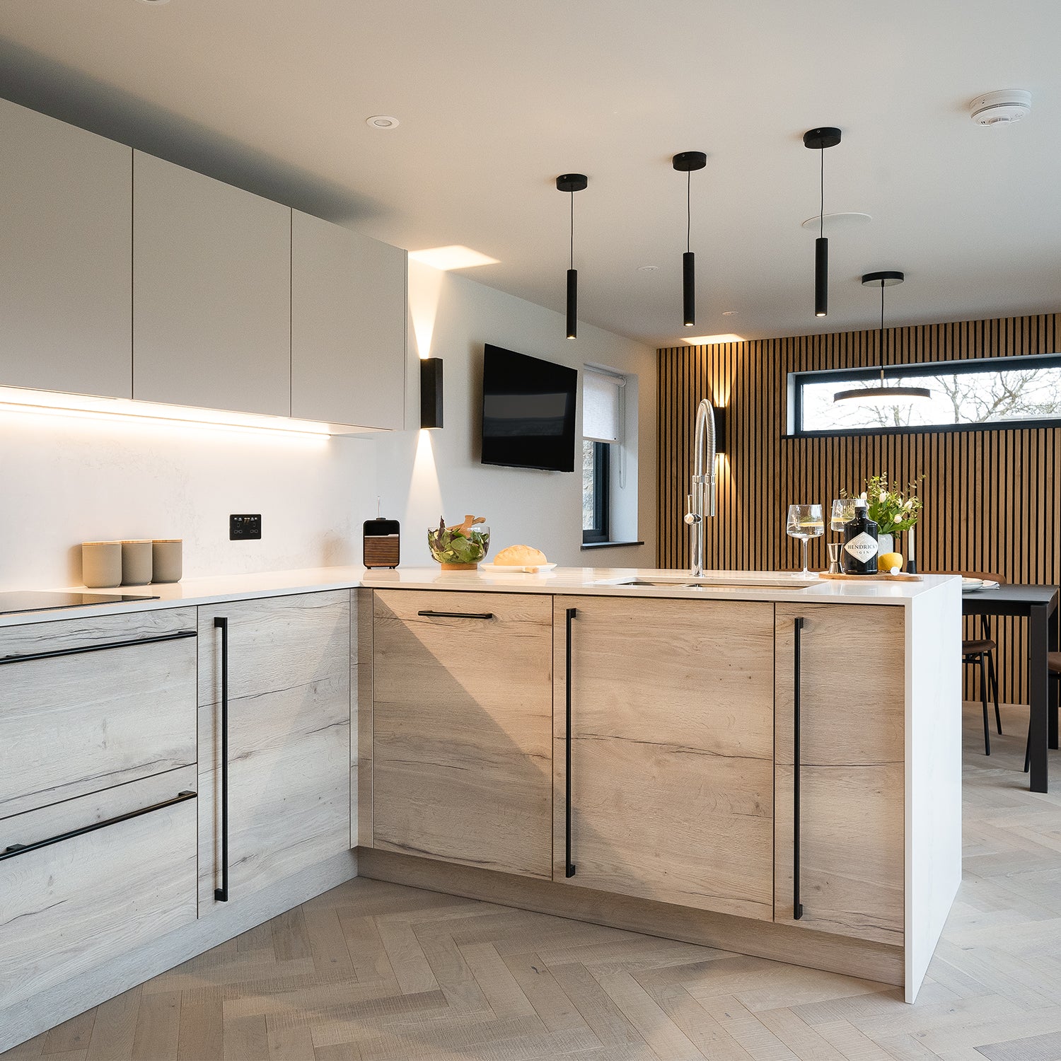 Contemporary Cornwall kitchen with wood-effect cabinets, white quartz worktops, matte black fixtures, herringbone flooring, and vertical slatted feature wall – modern kitchen design and bespoke cabinetry by interior design specialists in Cornwall.