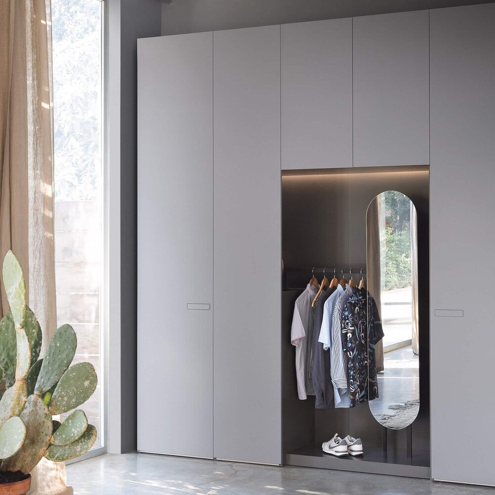 Minimalist built-in wardrobe with integrated open hanging space, soft grey doors, ambient lighting and full-length mirror – bespoke fitted furniture solution by Iroka Interiors, Cornwall, for modern and functional bedroom design.