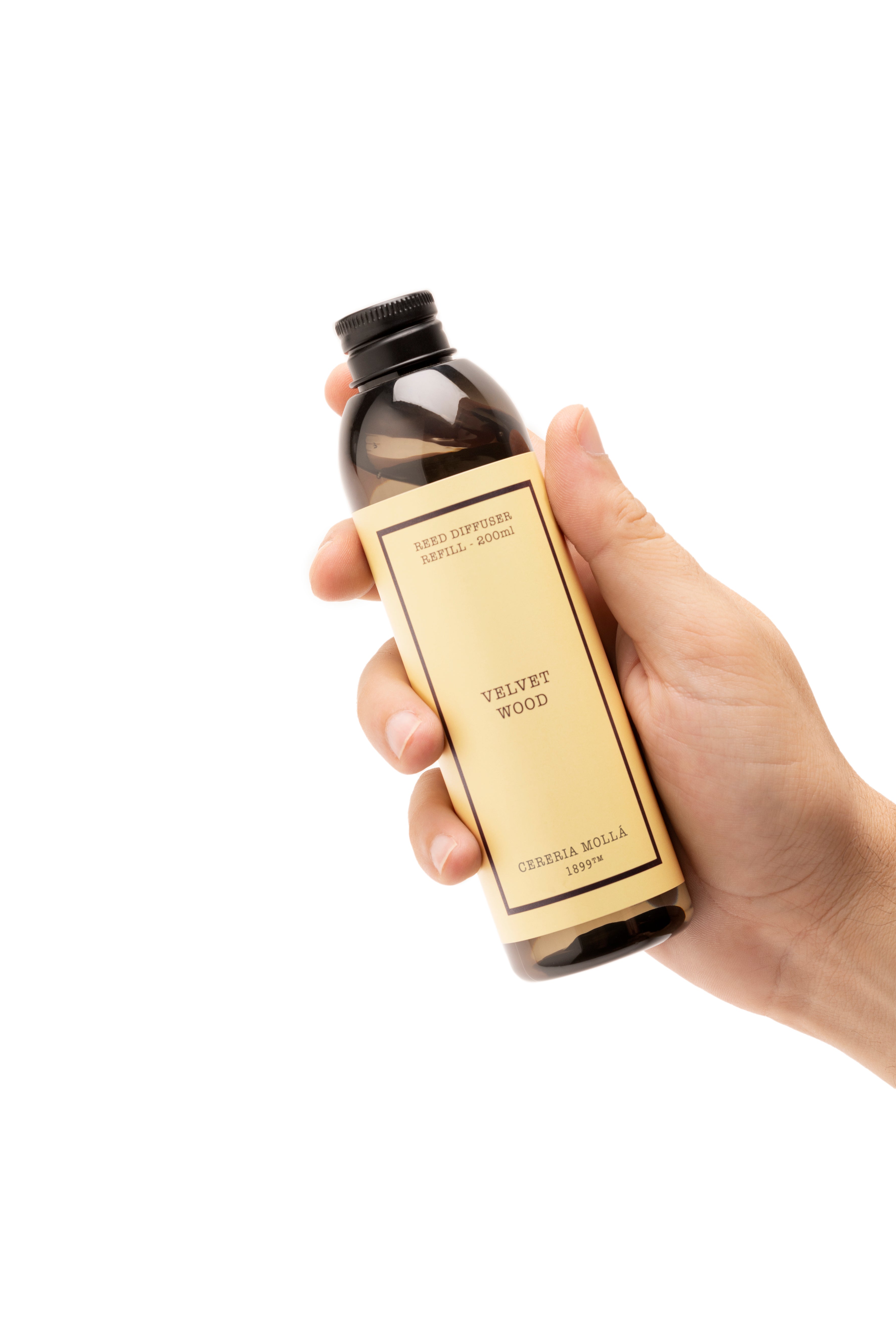 A hand holding a dark amber glass bottle with a black cap and a beige label that reads 'VELVET WOOD' and 'cereria mola' on a plain white background.