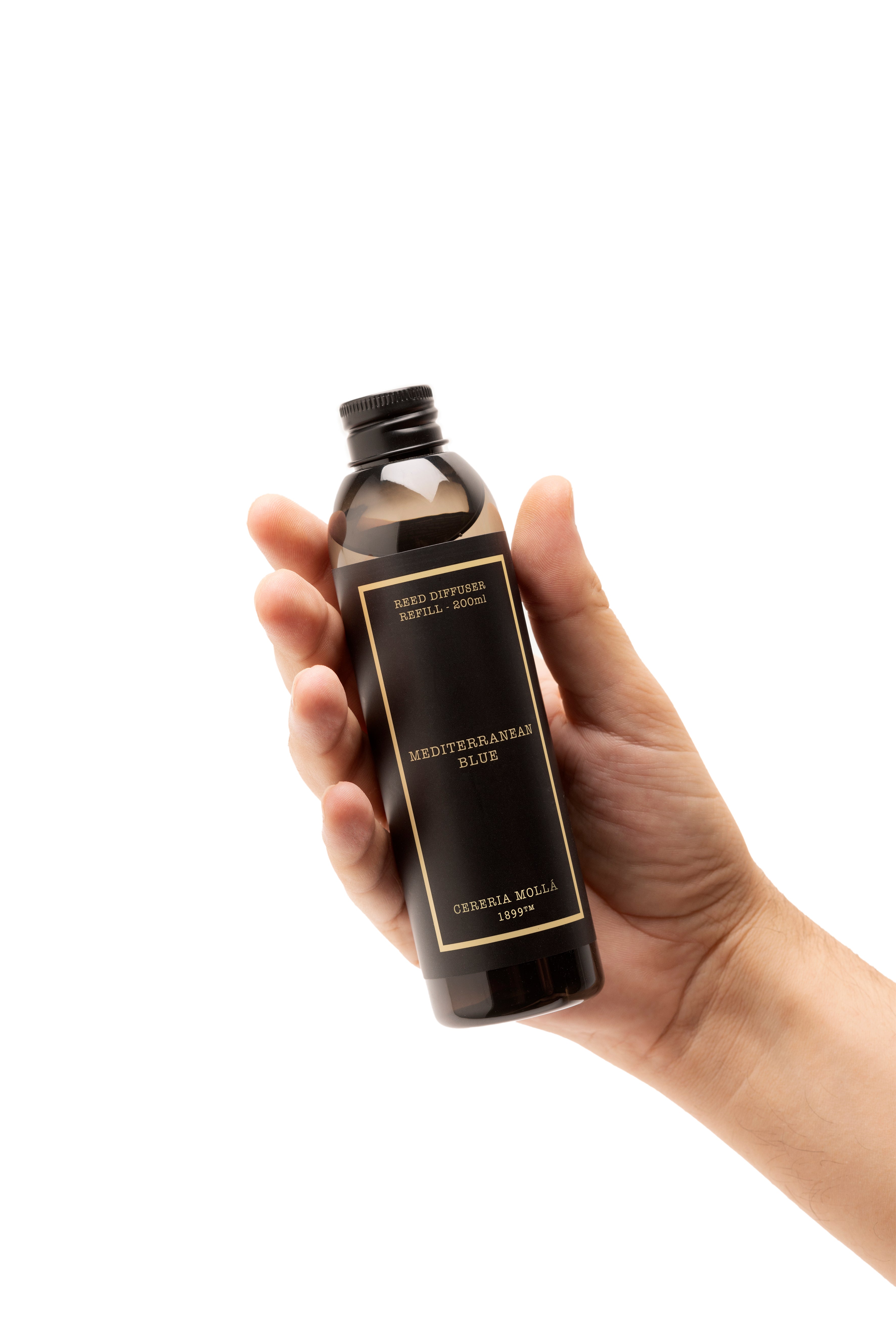 A hand holding a dark amber glass bottle with a black cap and a black label featuring gold lettering that reads 'MEDITERRANEAN blue' reed diffuser refill. The background is solid white.