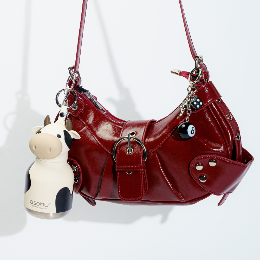 A glossy red shoulder bag with buckles, studs, and hanging charms, including a cow-shaped keychain labeled asobu, photographed against a plain white background.