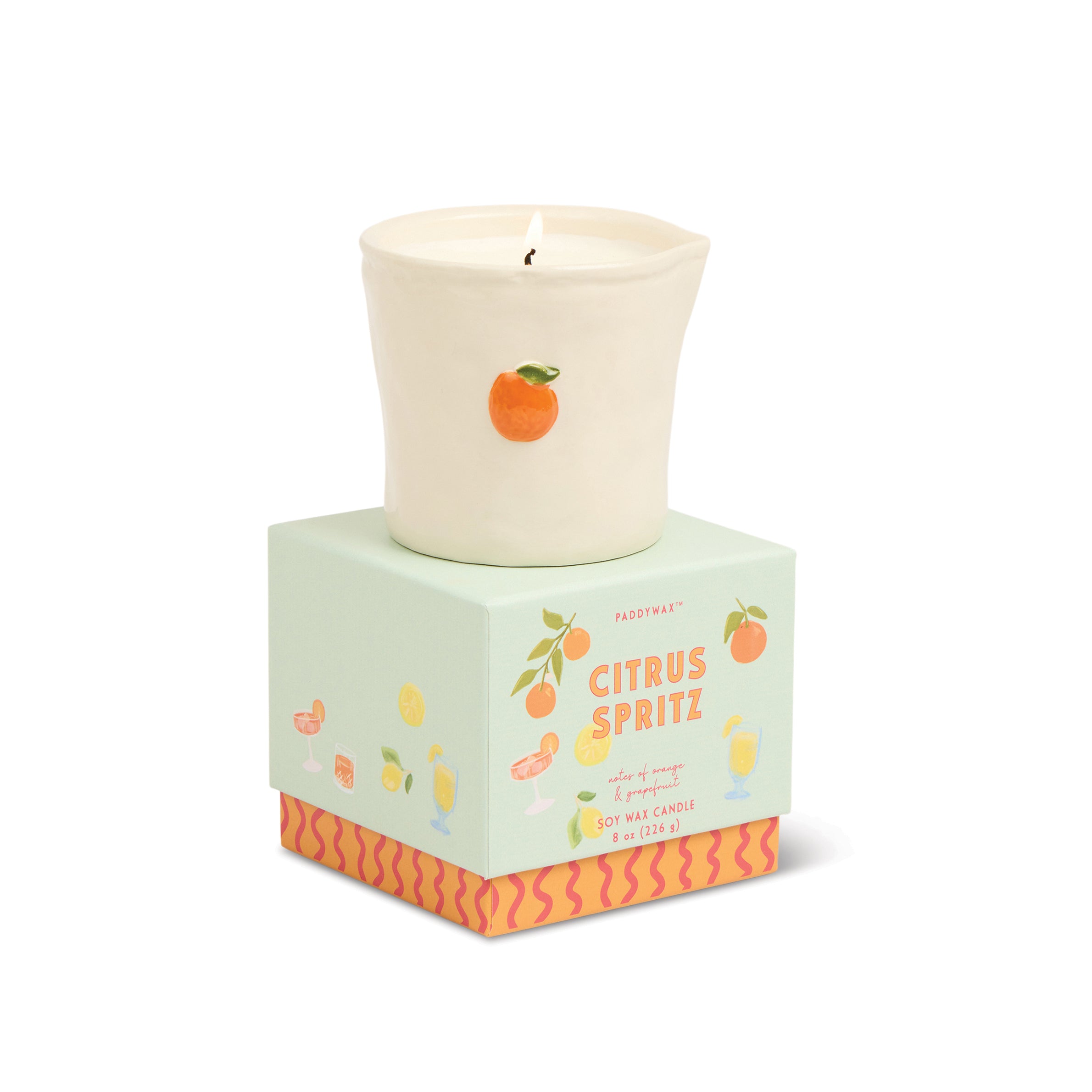 A lit cream-colored soy wax candle in a tapered cup, displayed on top of its mint green and orange patterned square packaging box. The box is labeled 'Citrus Spritz' and features illustrations of citrus fruits.
