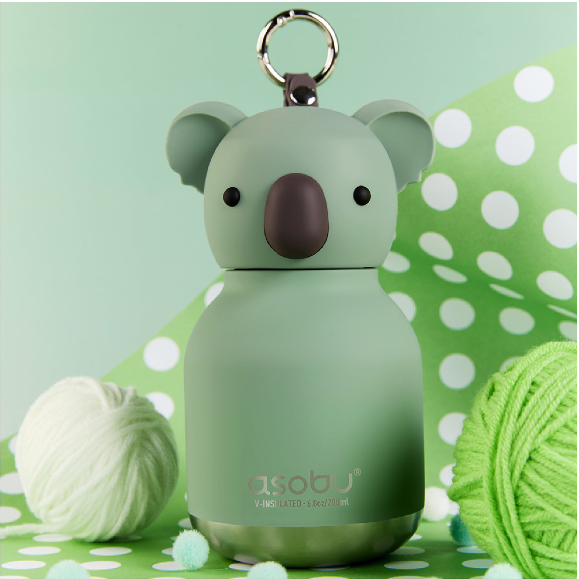 A cute koala-shaped water bottle by asobu in sage green, featuring a metal key ring at the top, set against a green and white polka dot background with decorative yarn balls.