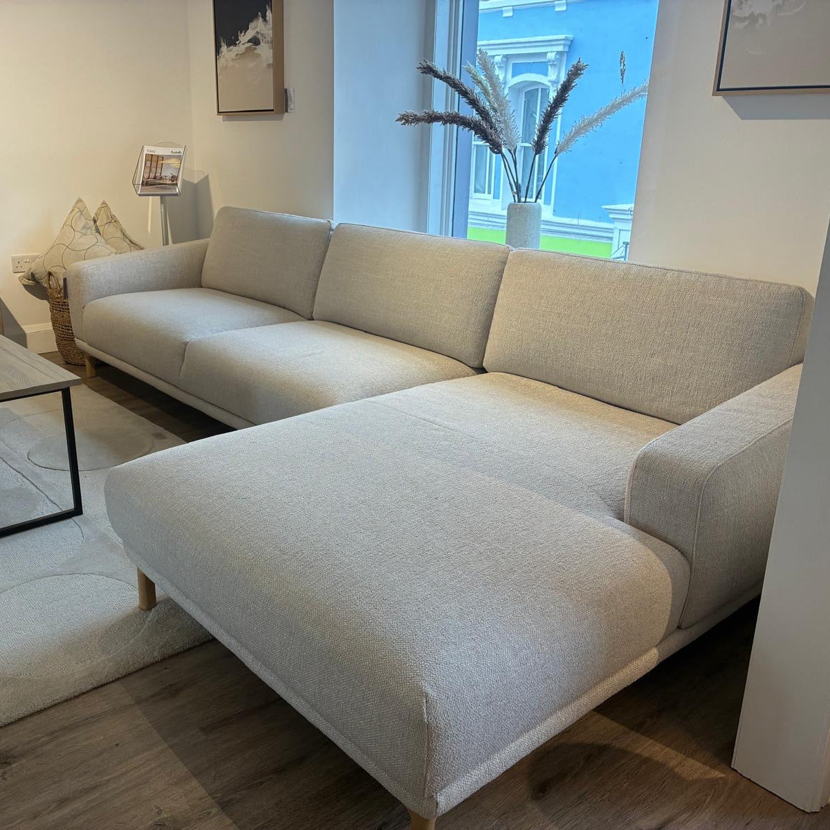 A light beige fabric chaise lounge sofa in a modern living room setting, positioned next to a matching light wood coffee table and a patterned rug.