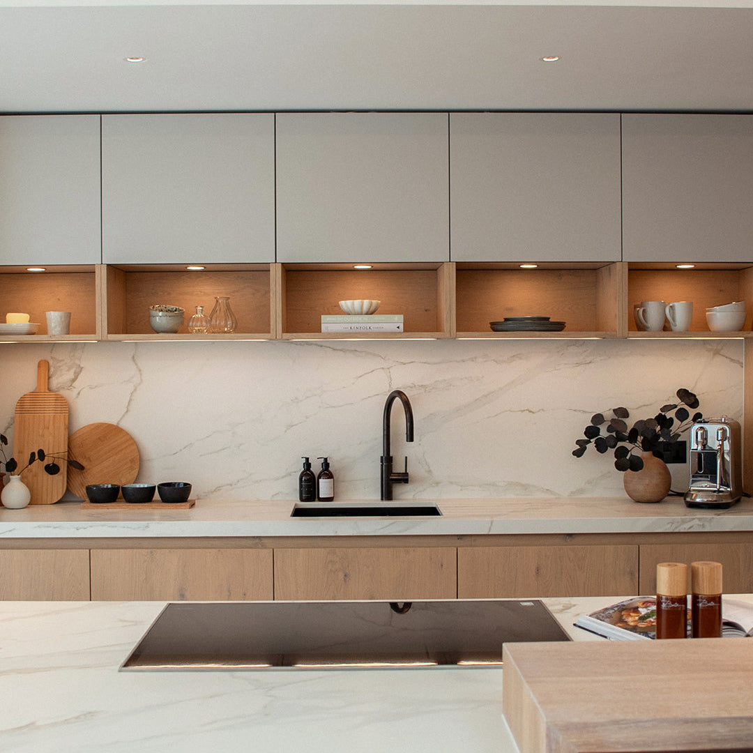 Modern kitchen interior with light gray upper cabinets, open wood shelves with small dishes, a white marble backsplash, a black faucet, and a light wood island in the foreground.