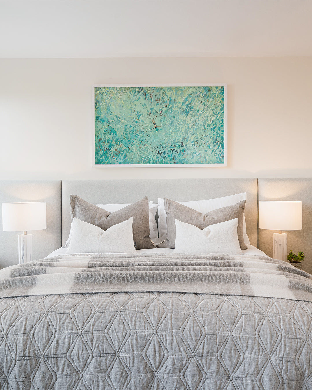 Calming coastal-inspired bedroom at Seaglass House, designed, supplied, and installed by Iroka Interiors. Features a soft grey upholstered headboard, layered neutral linens, sculptural bedside lamps, and a seaglass-toned aerial artwork - blending tranquility with tailored comfort.