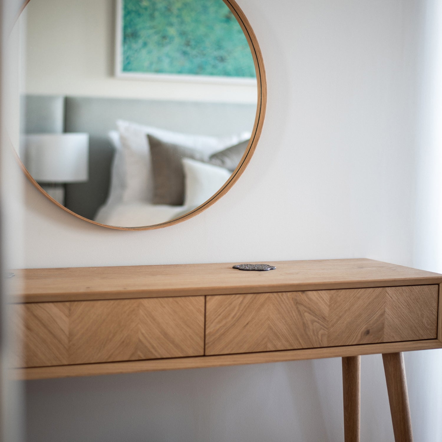 Detail from Clodgy View interior design project by Iroka Interiors. A circular oak-framed mirror reflects a serene bedroom with neutral linens and coastal artwork, paired with a natural wood chevron console table for a minimalist, coastal-inspired aesthetic.
