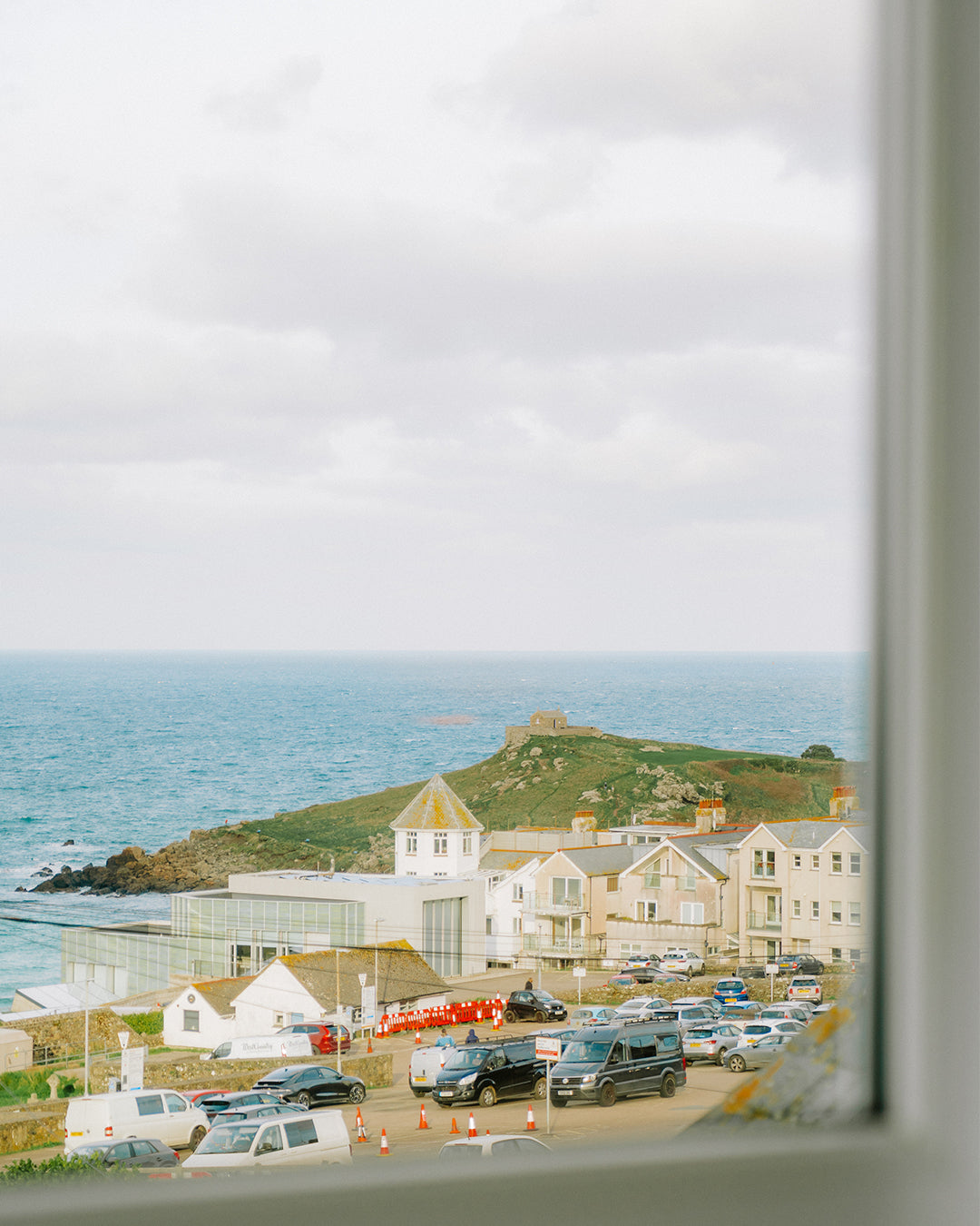 Scenic coastal view from Clodgy View in St Ives, Cornwall, featuring the Tate St Ives, historic seaside homes, and the Atlantic Ocean beyond, as seen through a window - capturing the charm of this Cornish coastal retreat.