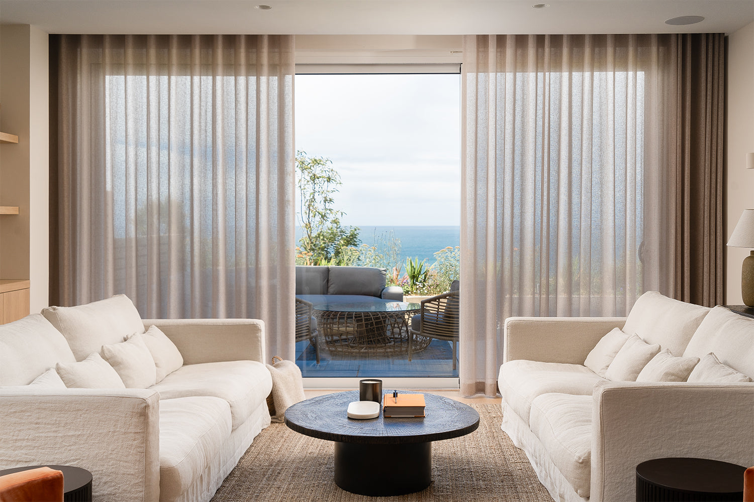 Modern living room with white sofas, a coffee table, and large windows overlooking an ocean view.