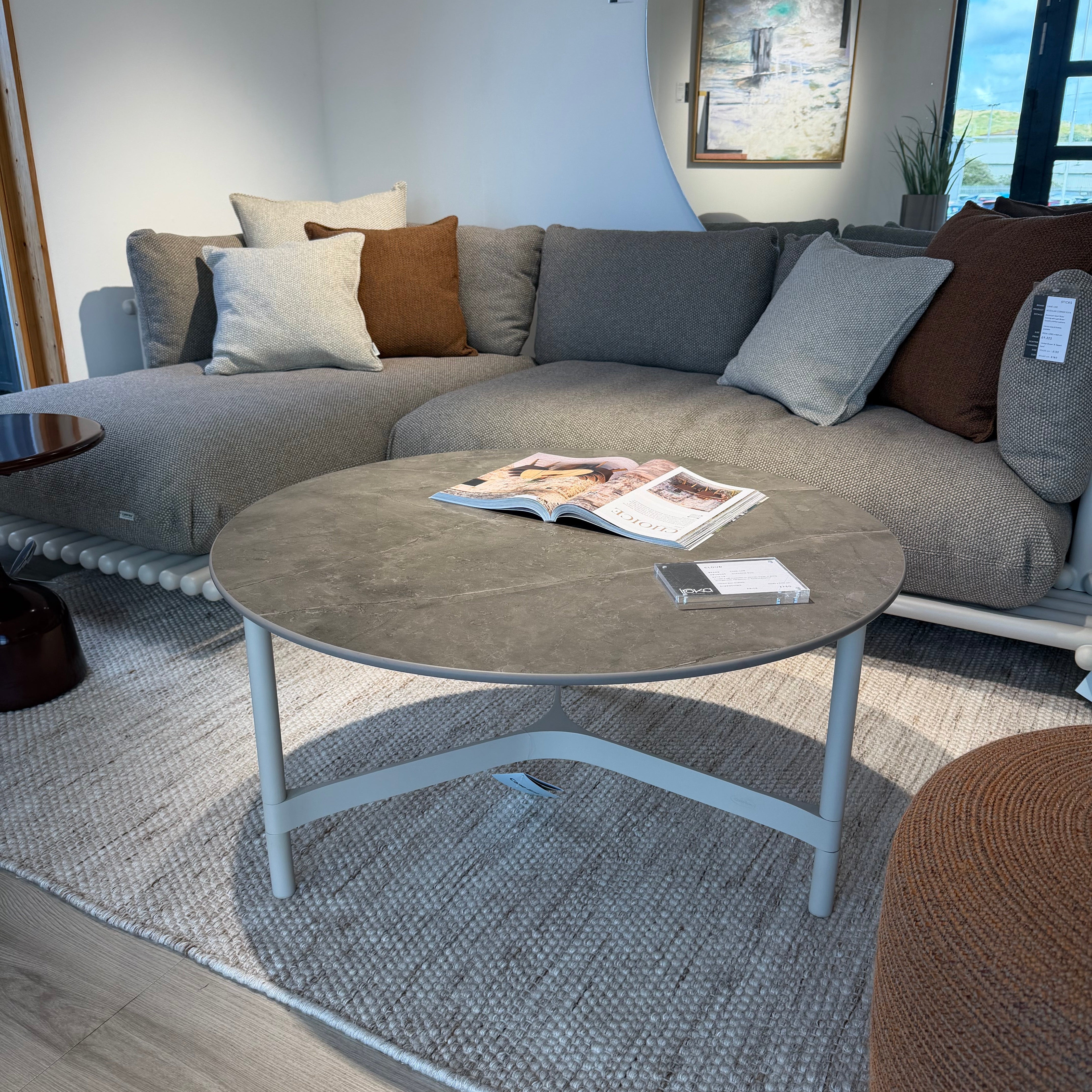 A modern gray sectional sofa with neutral and brown accent pillows, styled in a bright living room with a round gray coffee table, textured rug, and light wood flooring.