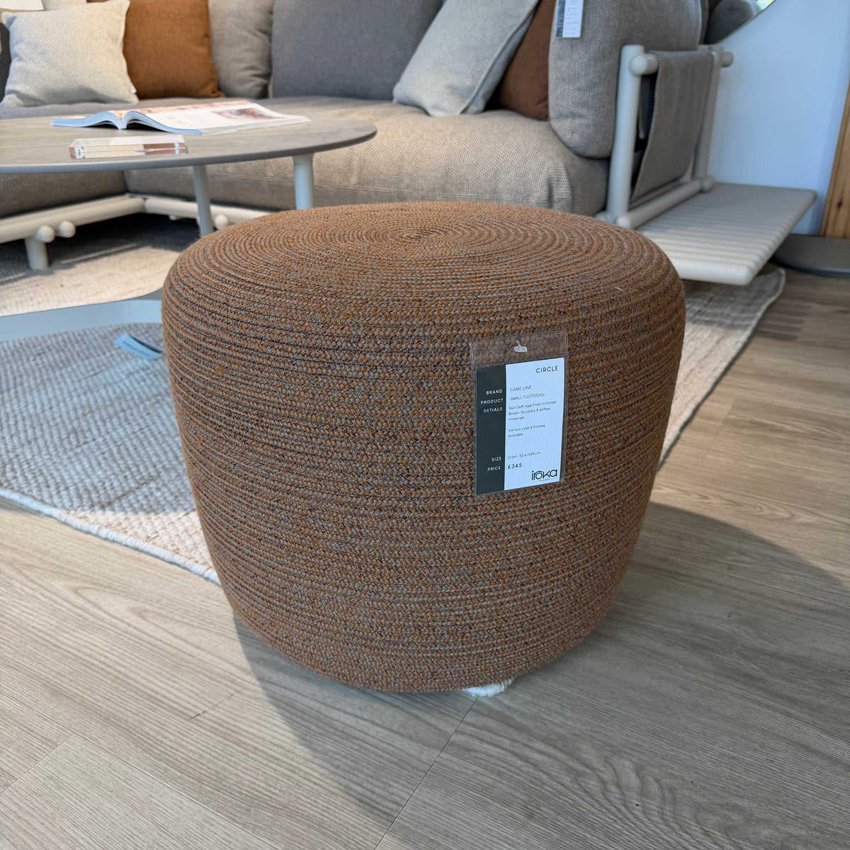 A round, ribbed brown upholstered ottoman with a white product tag attached, set on a light-colored rug in a modern living room environment.