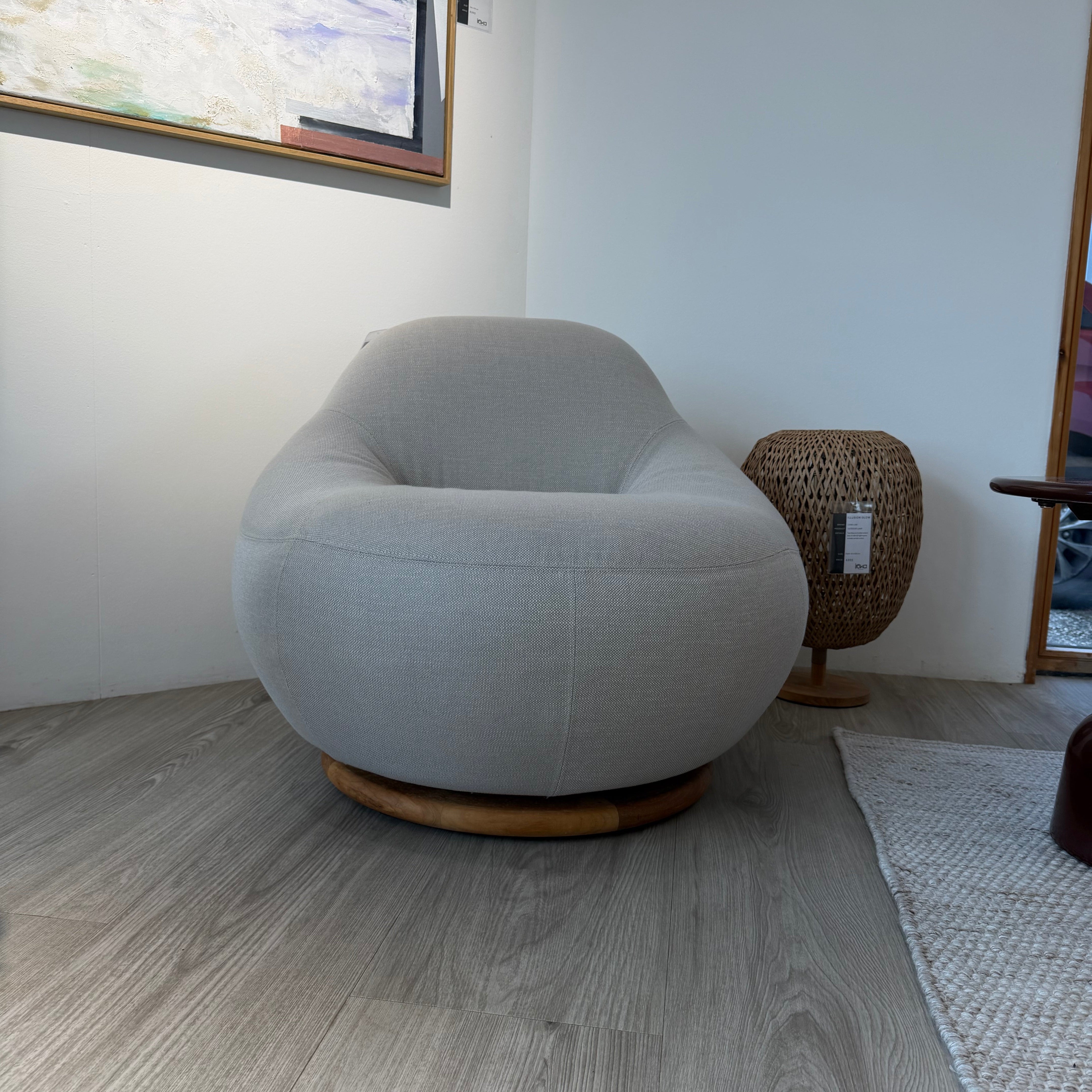 A modern round swivel armchair upholstered in light gray textured fabric with a natural wood base, shown in a minimalist room setting with gray wood-look flooring and a framed painting on the wall.