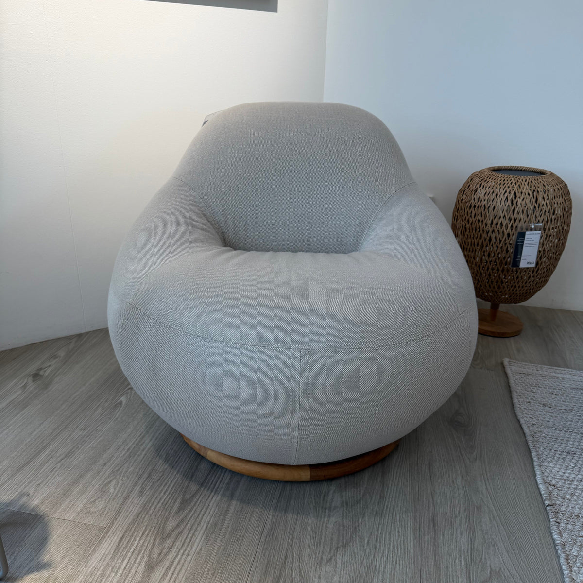 A light gray upholstered swivel armchair with a round wooden base, photographed in a minimalist room with gray wood-look flooring and a woven side table nearby.
