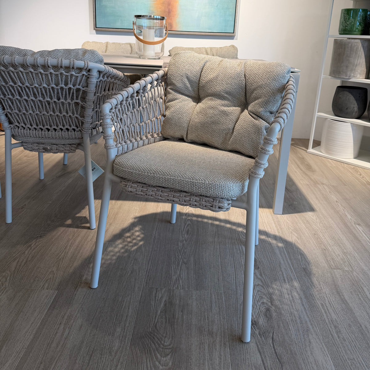 A modern outdoor dining chair with woven gray wicker and a tufted gray cushion, shown in a staged indoor room with wood flooring, a dining table, and wall art.