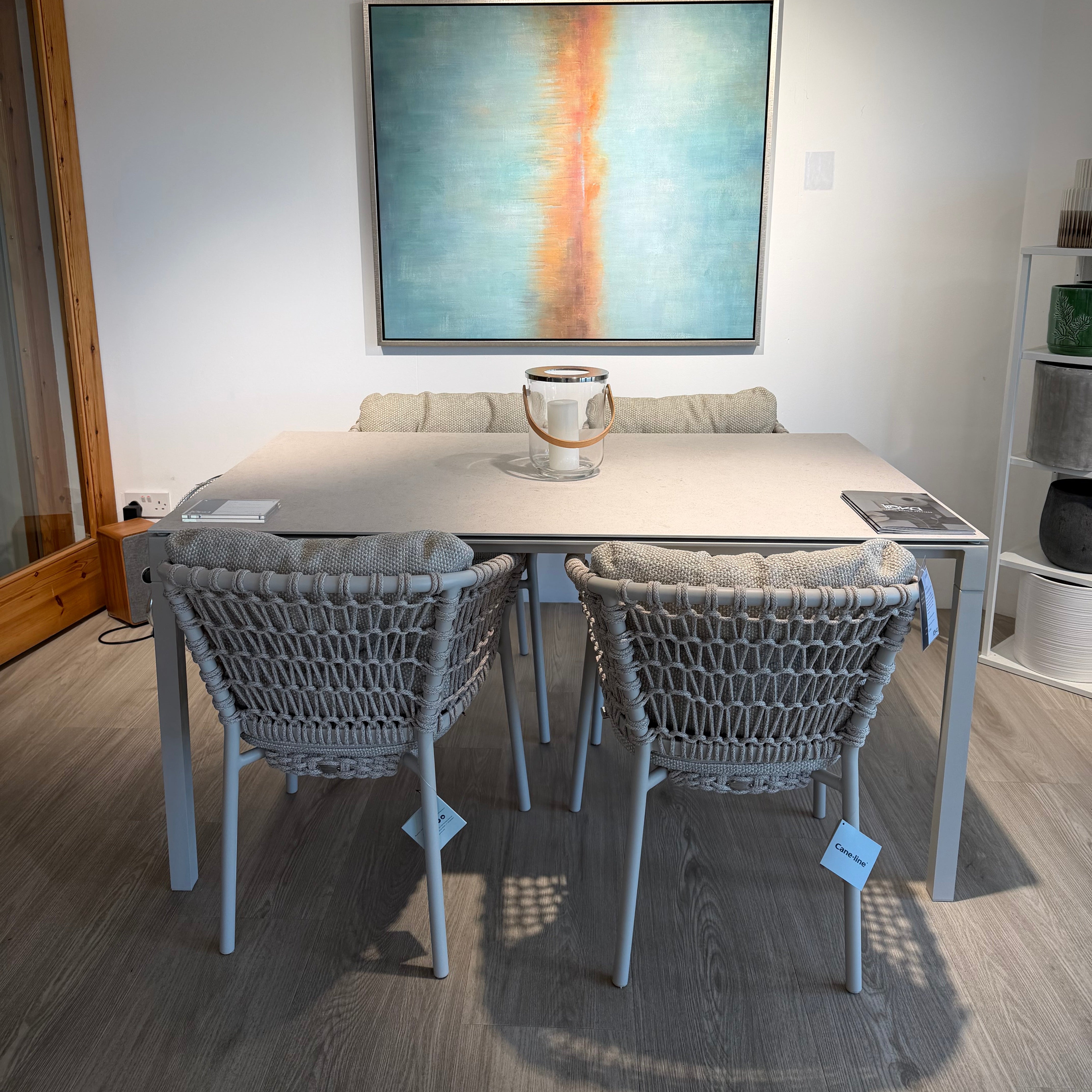 A modern outdoor dining set consisting of a rectangular light gray table and four woven gray chairs, displayed in a showroom with a large abstract painting on the wall behind it.