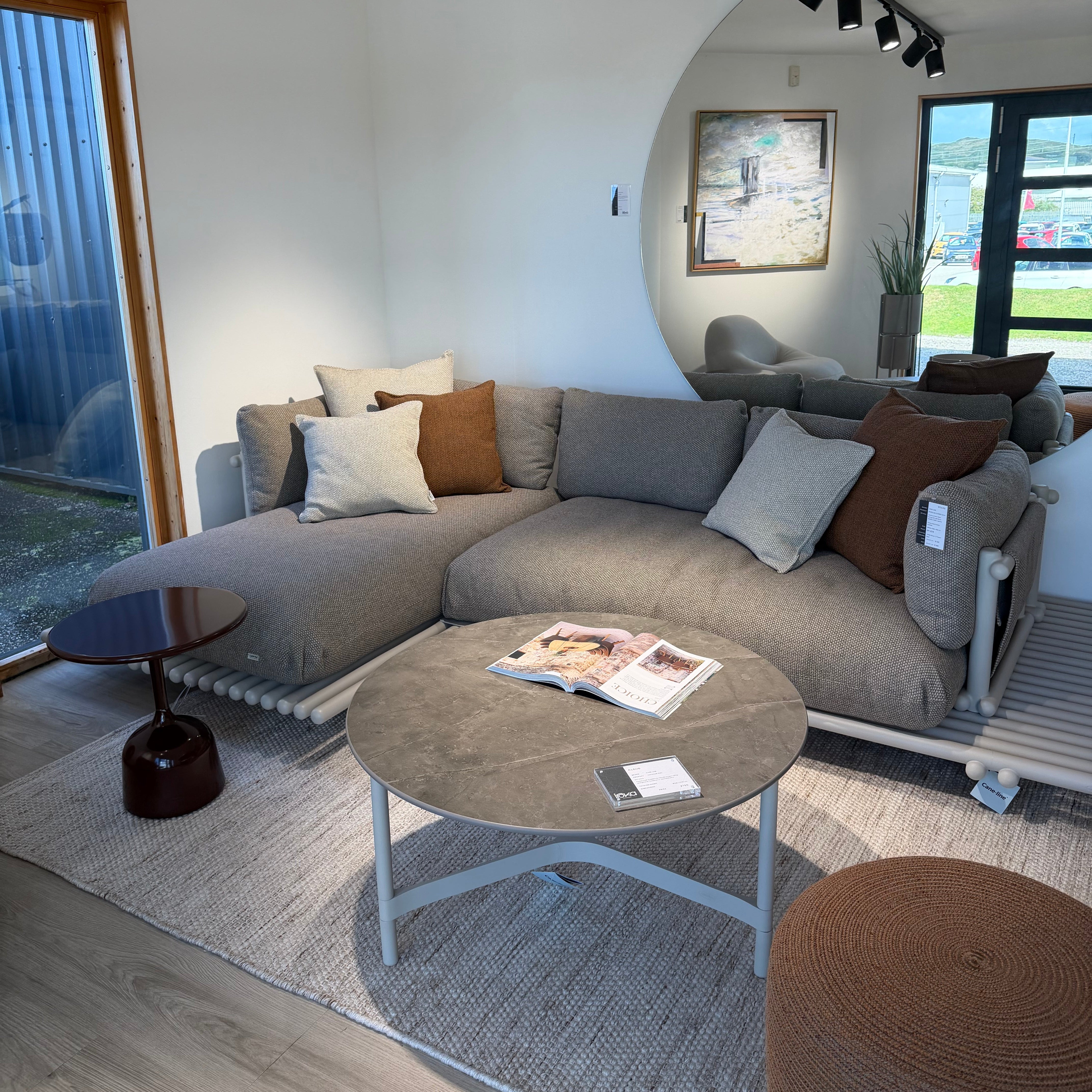A modern gray fabric sectional sofa with white and brown accent pillows, styled in a contemporary living room with a round coffee table, textured rug, and large arched window view.