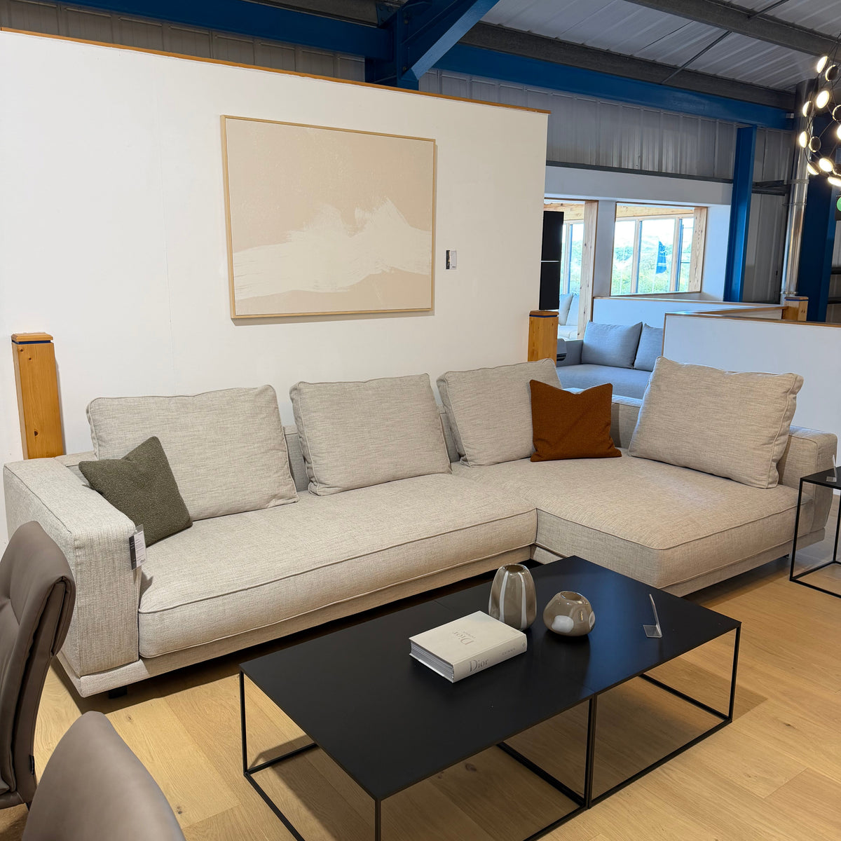 A modern light beige sectional sofa in a showroom setting, styled with neutral and brown accent pillows, a black coffee table, and a minimalist abstract painting on the wall.
